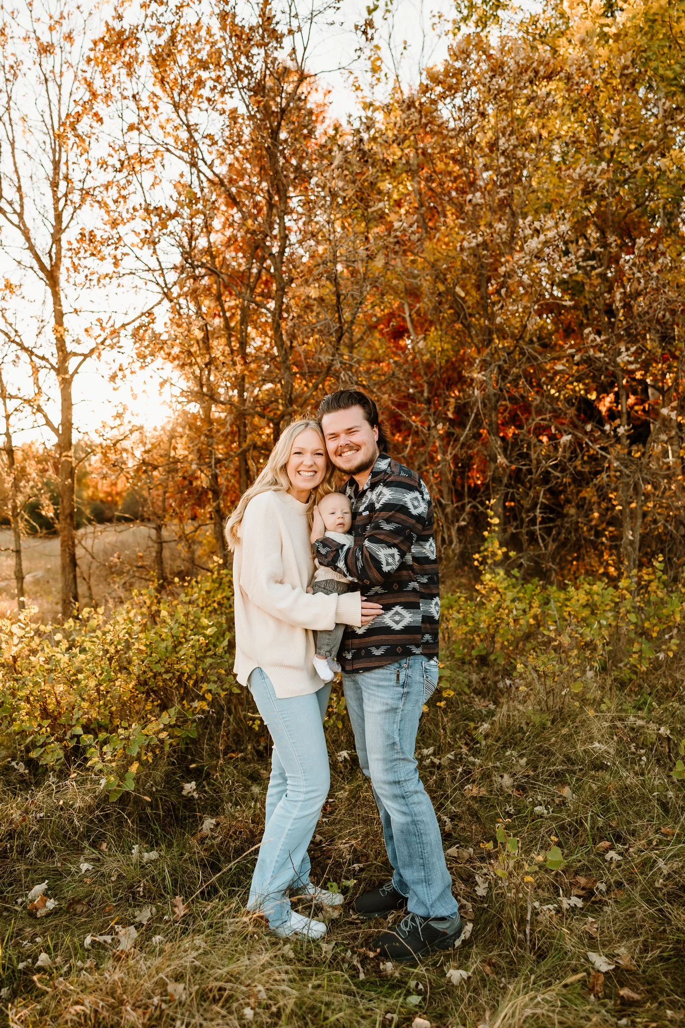 A happy family of three, including a woman, man, and baby, standing outdoors on autumn grass with colorful fall trees and a setting sun in the background.