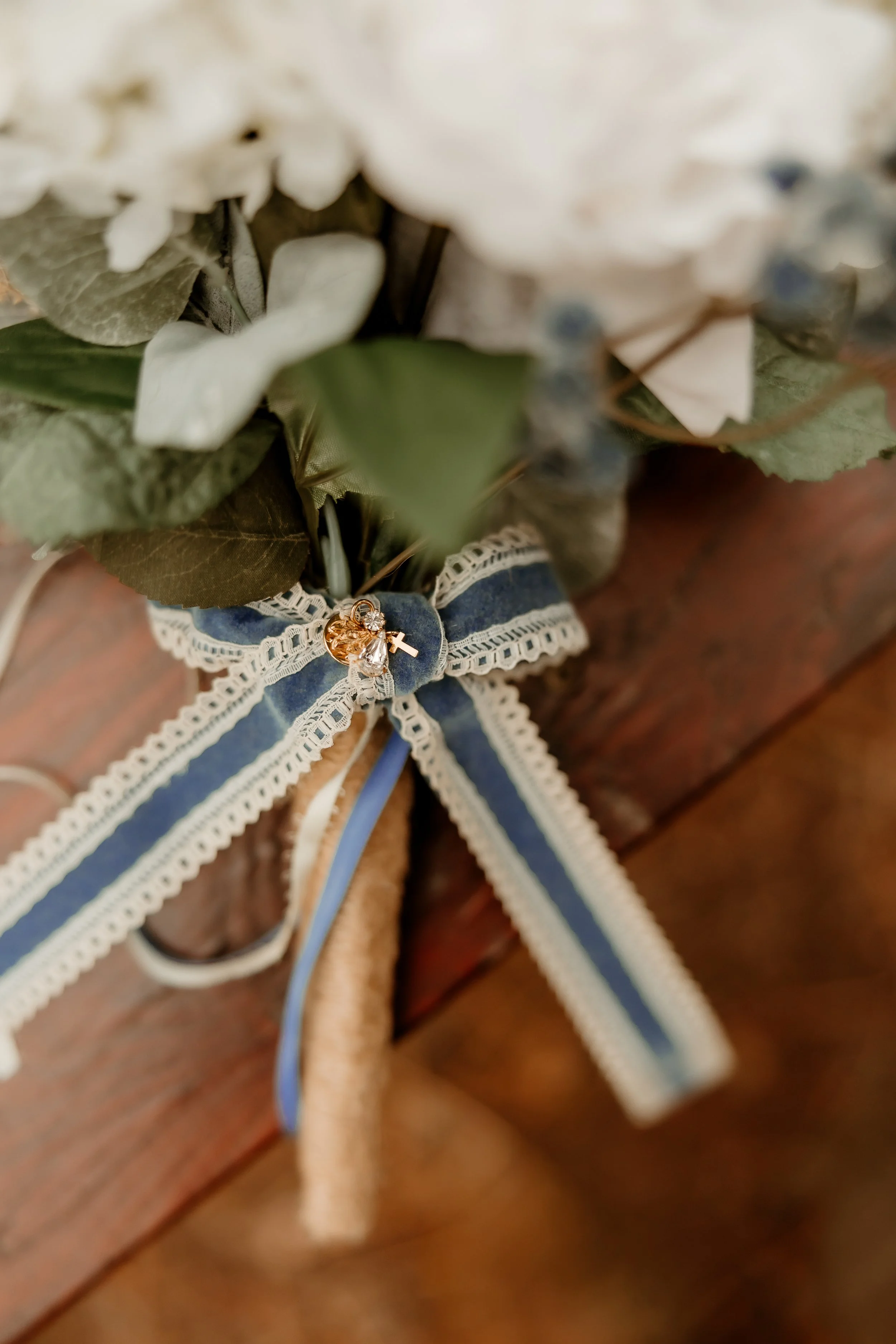 Close-up of a bouquet of flowers with a navy blue and white ribbon tied around the stems, decorated with small charms including a cross, a heart, and a clear gem, on a wooden surface.