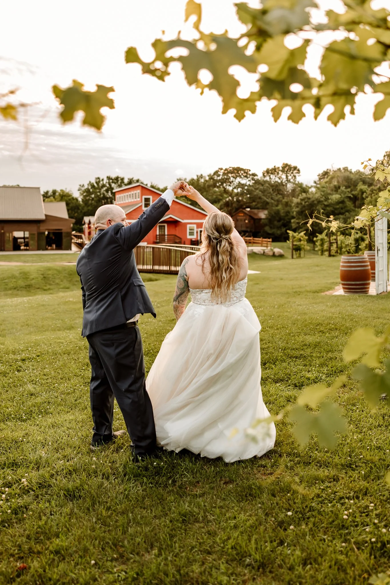A newlywed couple, dressed in wedding attire, dancing in a grassy outdoor setting during sunset with trees and colorful barns in the background.