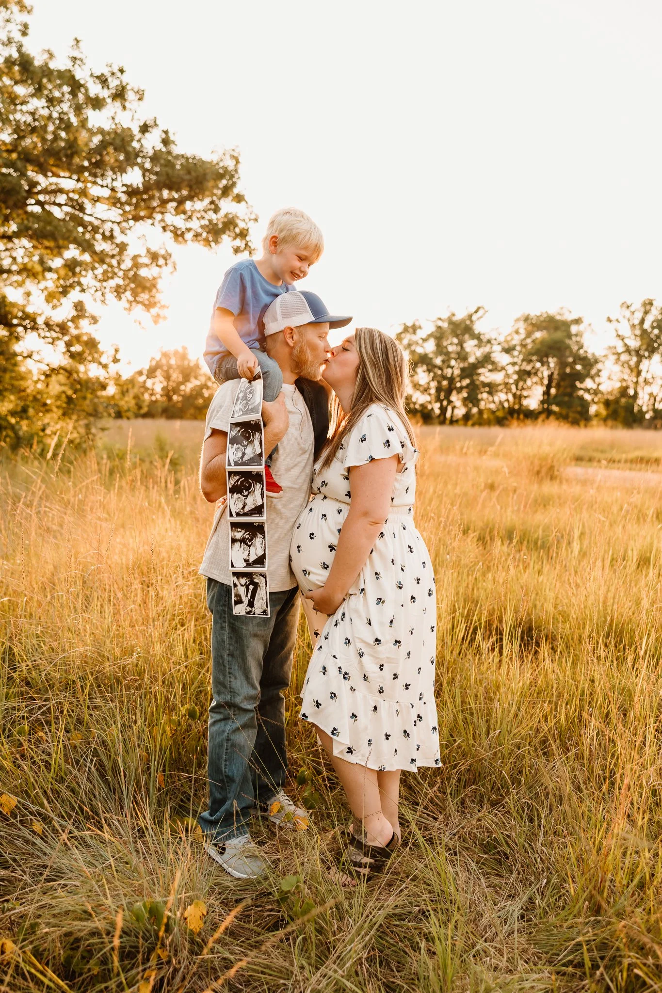 A family of three celebrating pregnancy outdoors during sunset. The father, with a beard and cap, holds their young son on his shoulders as they kiss the pregnant mother, all standing in a grassy field.