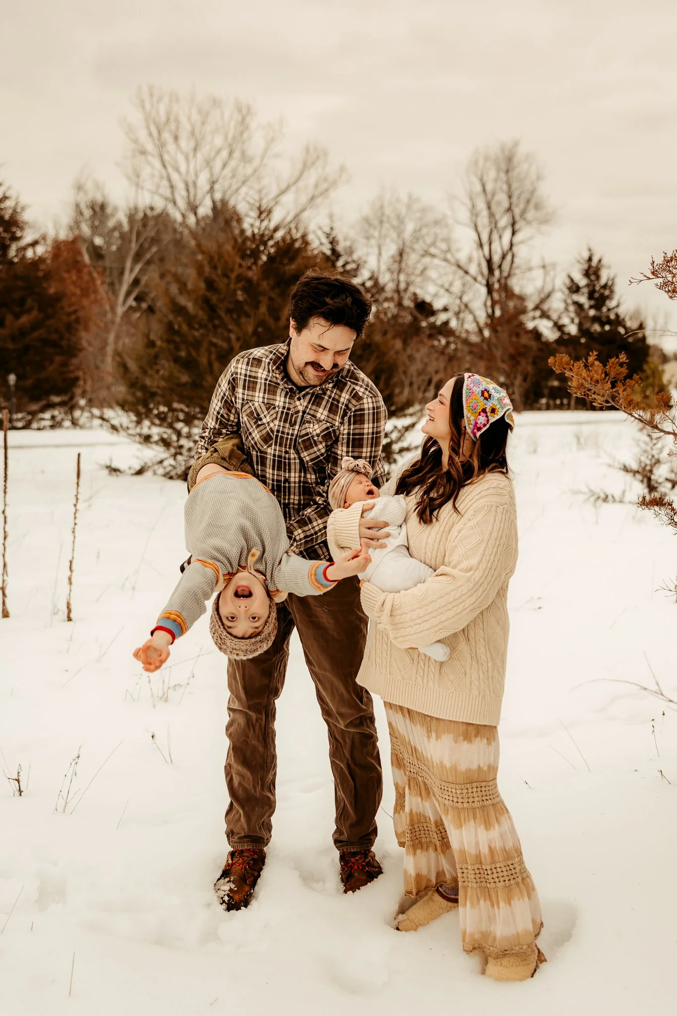 Family outdoors in snow, with man holding smiling toddler upside down, woman holding crying baby, all dressed warmly.