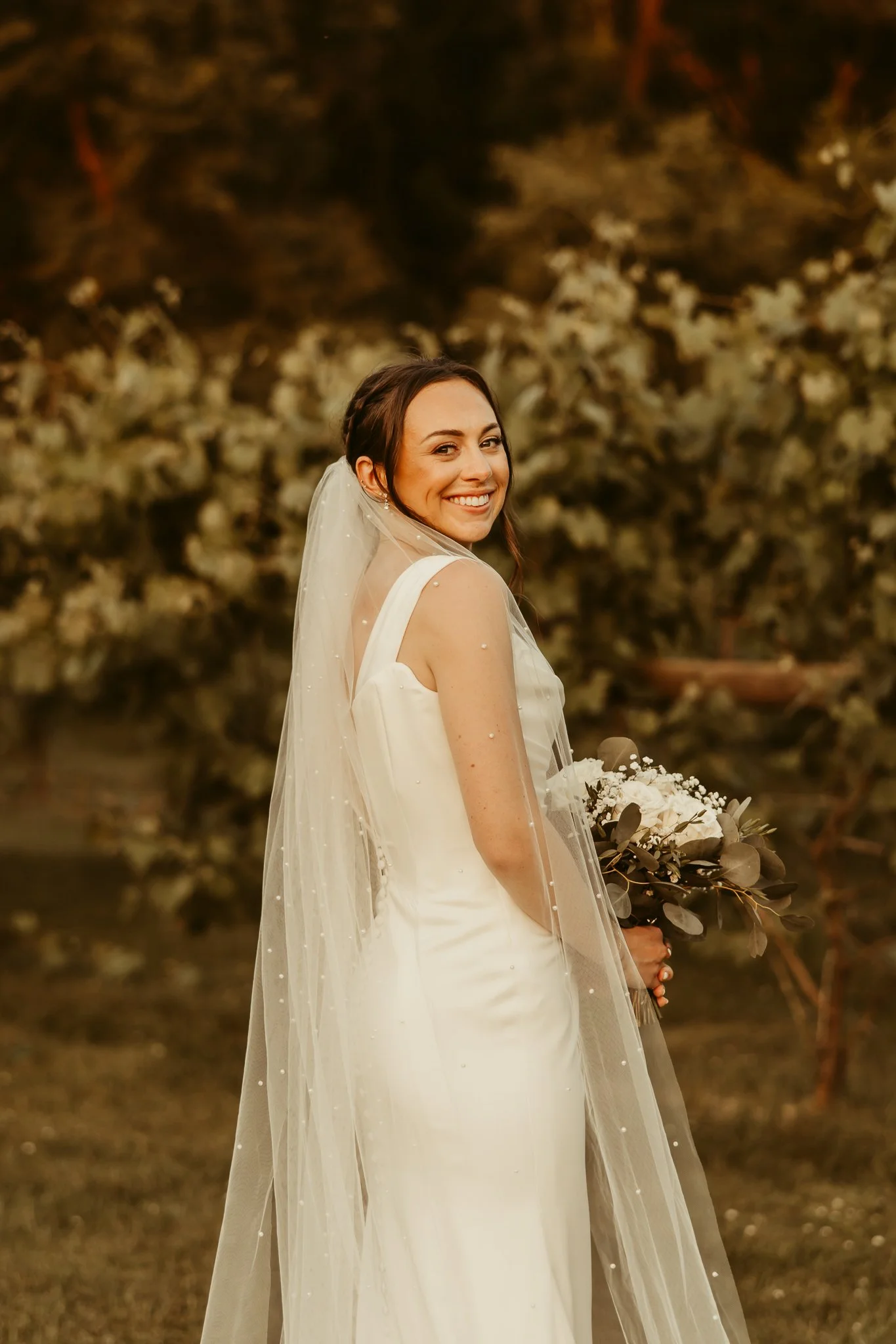A smiling bride in a white wedding dress holding a bouquet, standing outdoors with trees in the background.