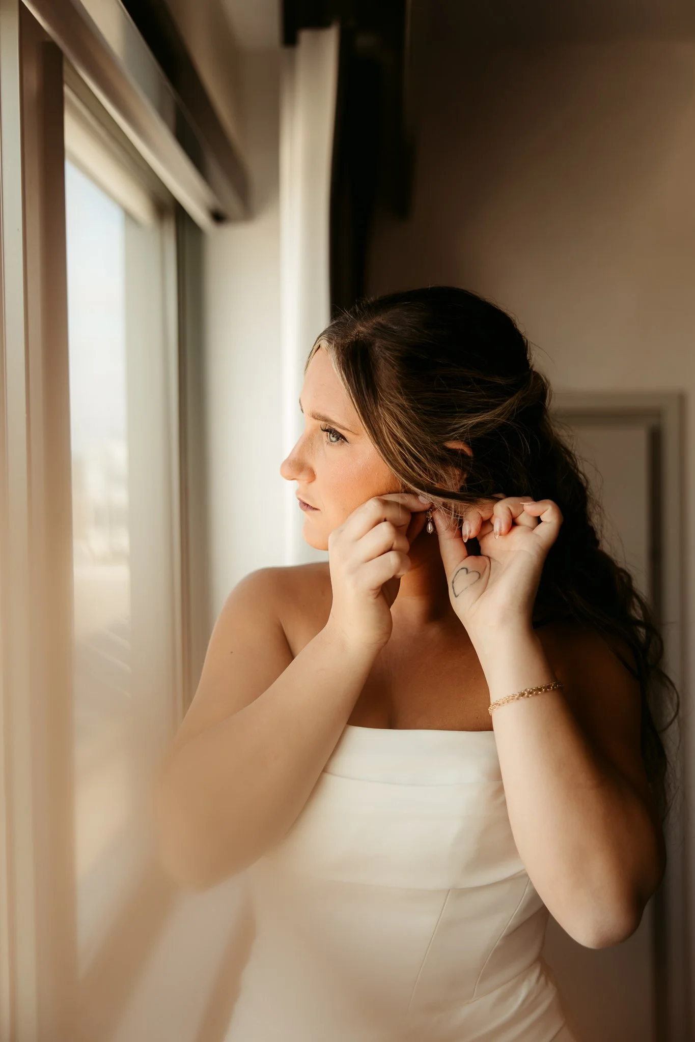A woman in a white dress is putting on an earring while looking out a window, with sunlight illuminating her face.