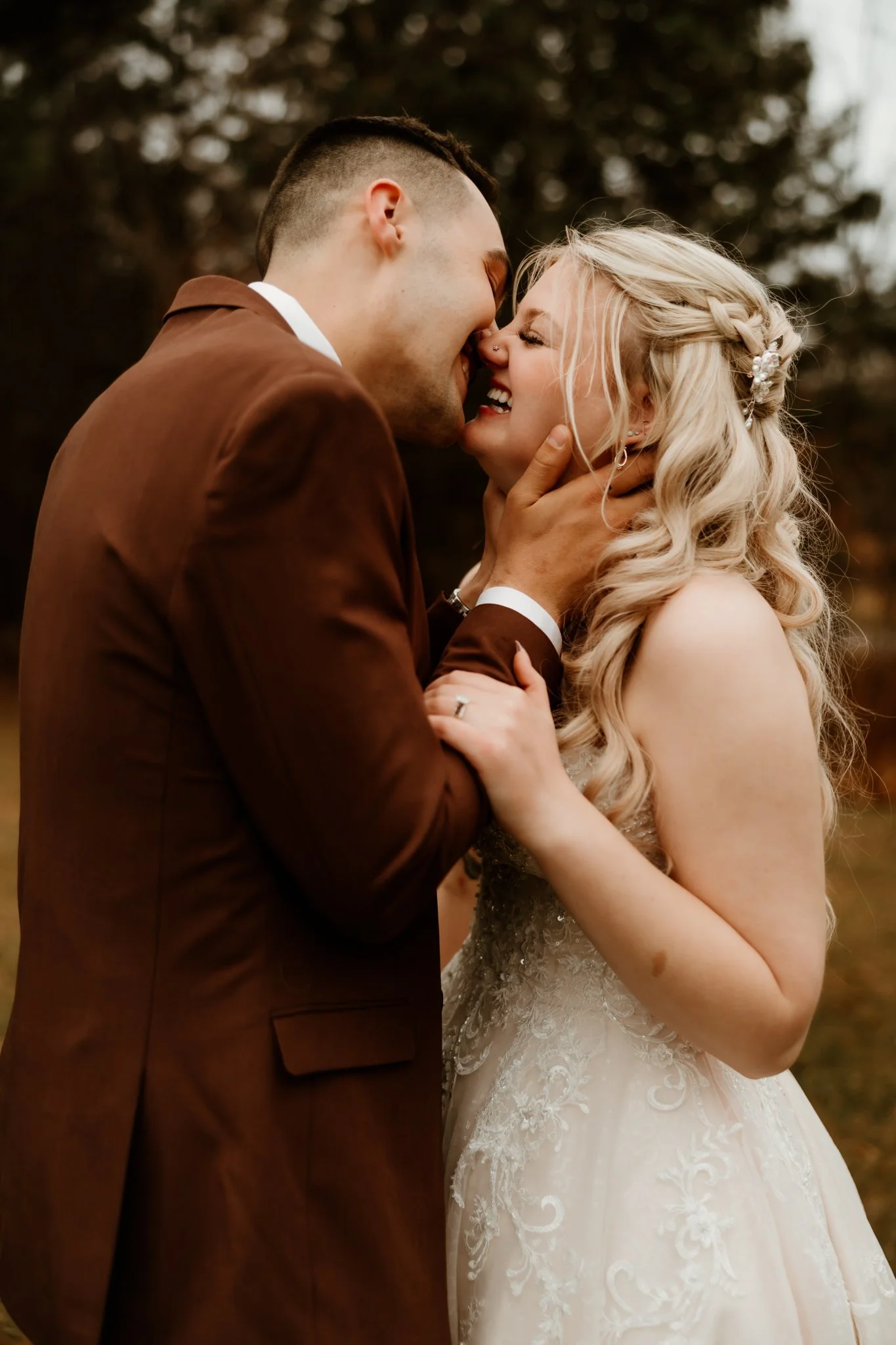 A couple, dressed in wedding attire, sharing a joyful moment outdoors with a blurred natural background.