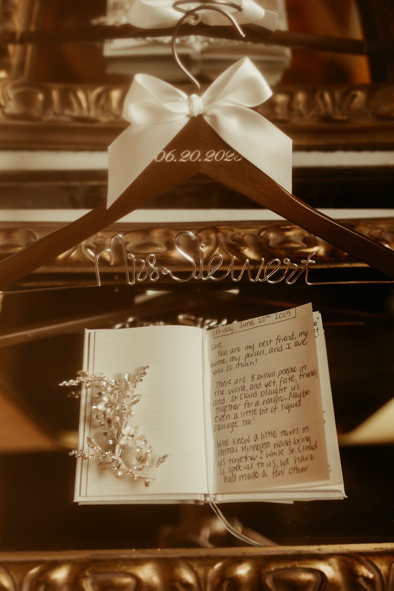 A wedding guestbook display with a wooden wedding hanger inscribed with the date 06.20.2023, a wire hanger with the name Miss Stewart, an open handwritten guestbook, and a decorative hairpiece resting on the guestbook. The setup is on a reflective su