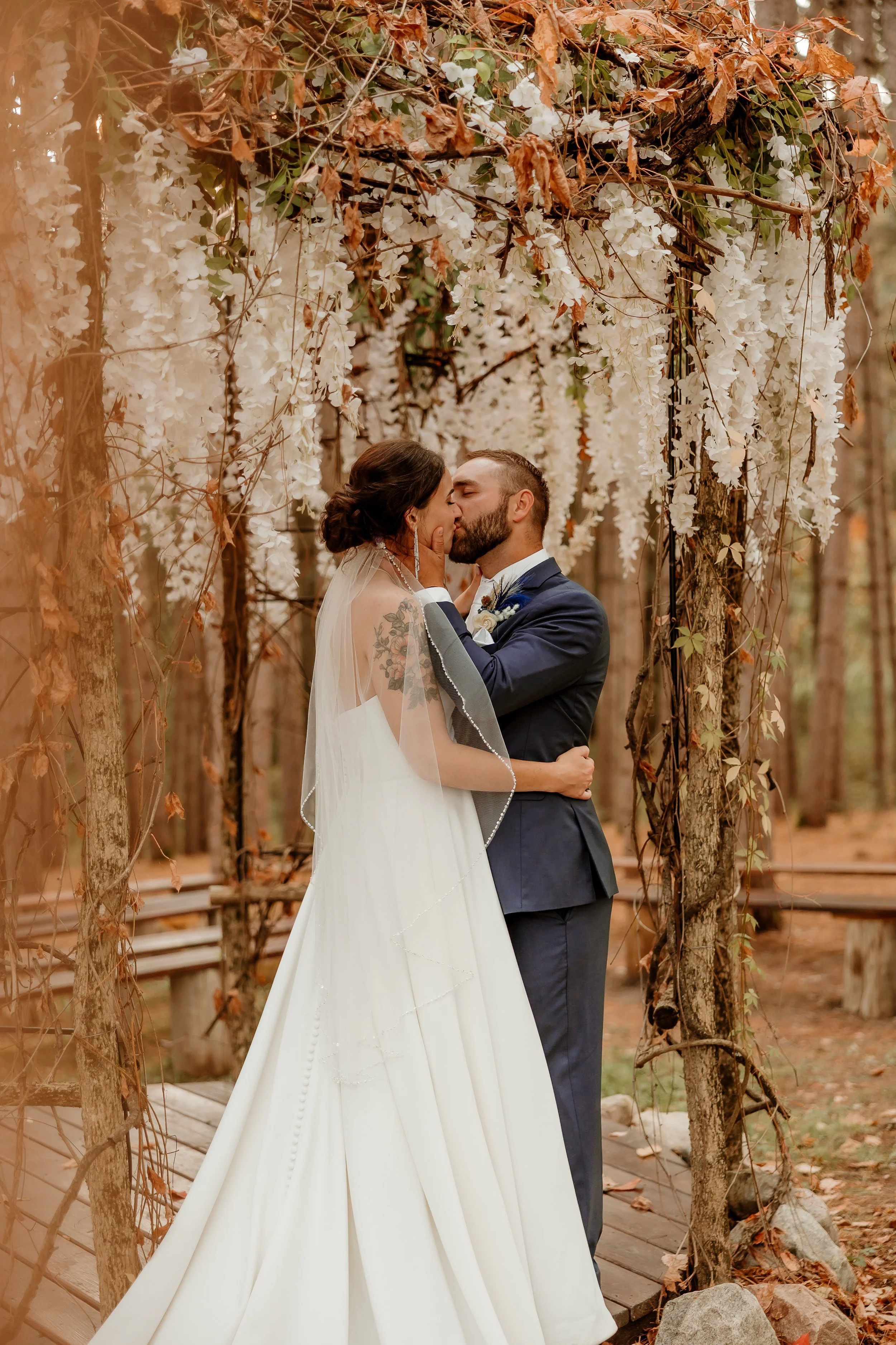 A bride and groom share a kiss under a floral arch in a forest setting.