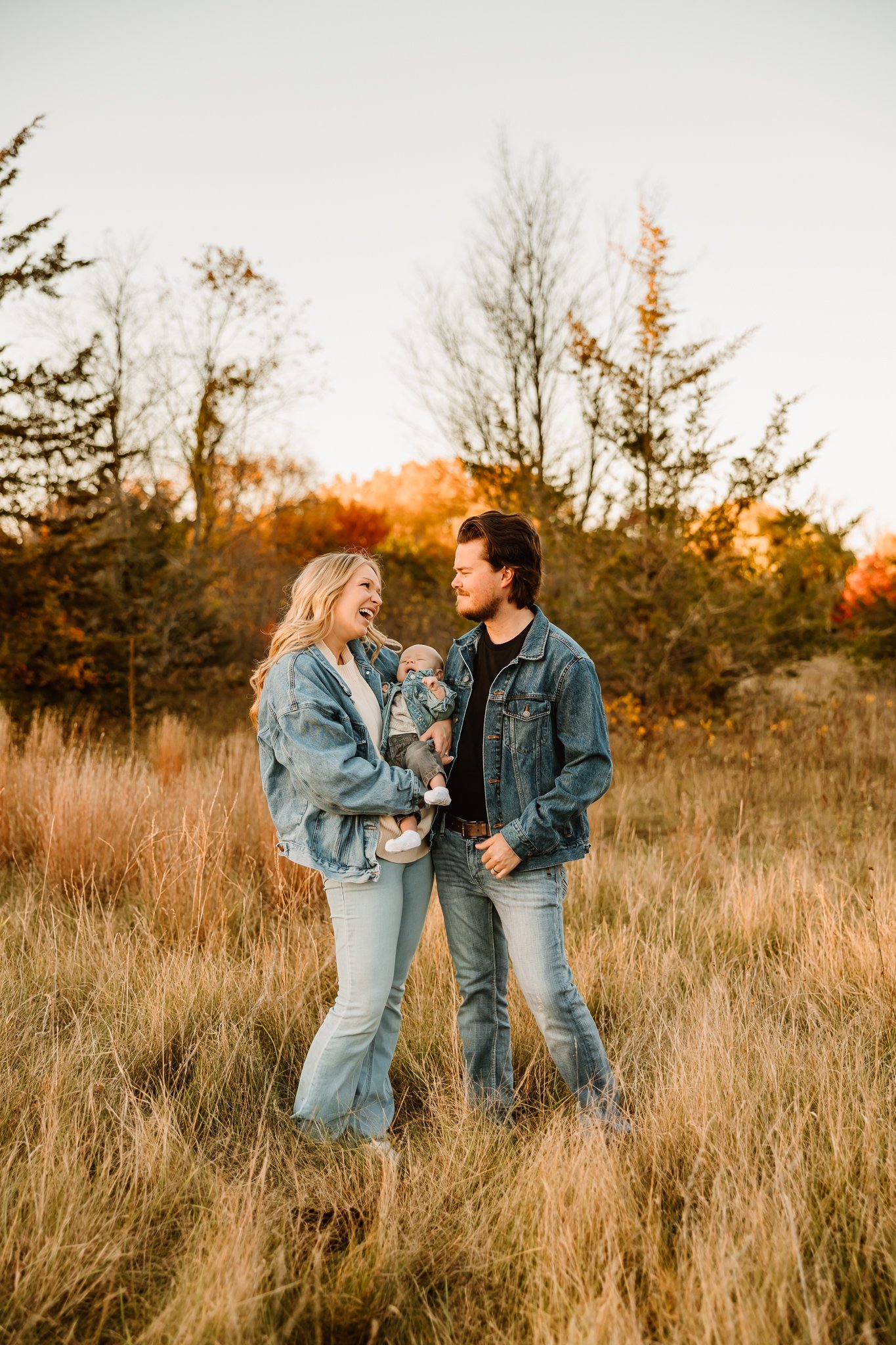A family of three, woman, man, and baby, standing in a grassy field with fall foliage in the background, smiling and enjoying time together.