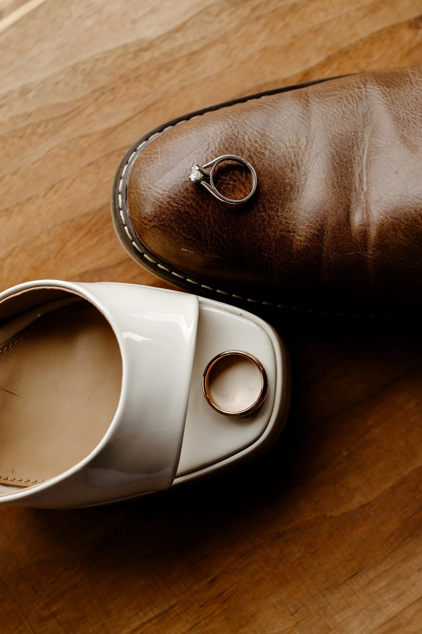 A white slip-on shoe with a ring on top, a brown leather boot with a ring on top, both placed on a wooden surface.