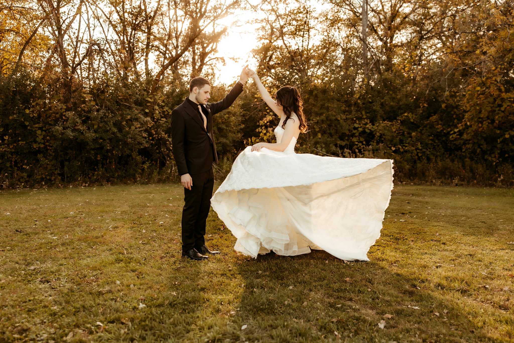 A couple in wedding attire dancing outdoors during sunset, with the groom in a black suit and the bride in a white wedding gown, on a grassy field surrounded by trees.