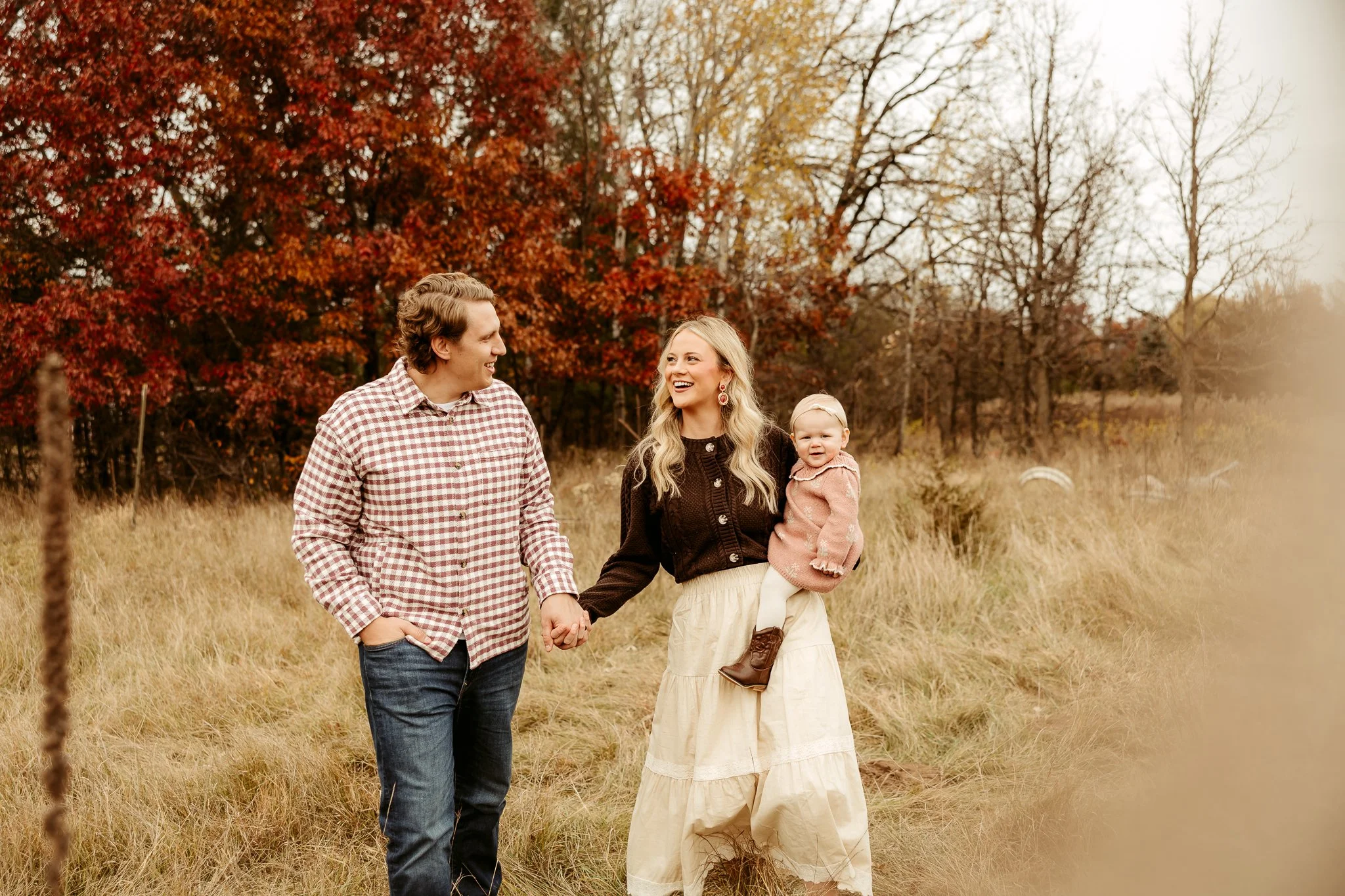 A family of three walking outdoors in a field during autumn, holding hands and smiling. The woman is carrying a young girl in her arm, and they are surrounded by trees with fall foliage.