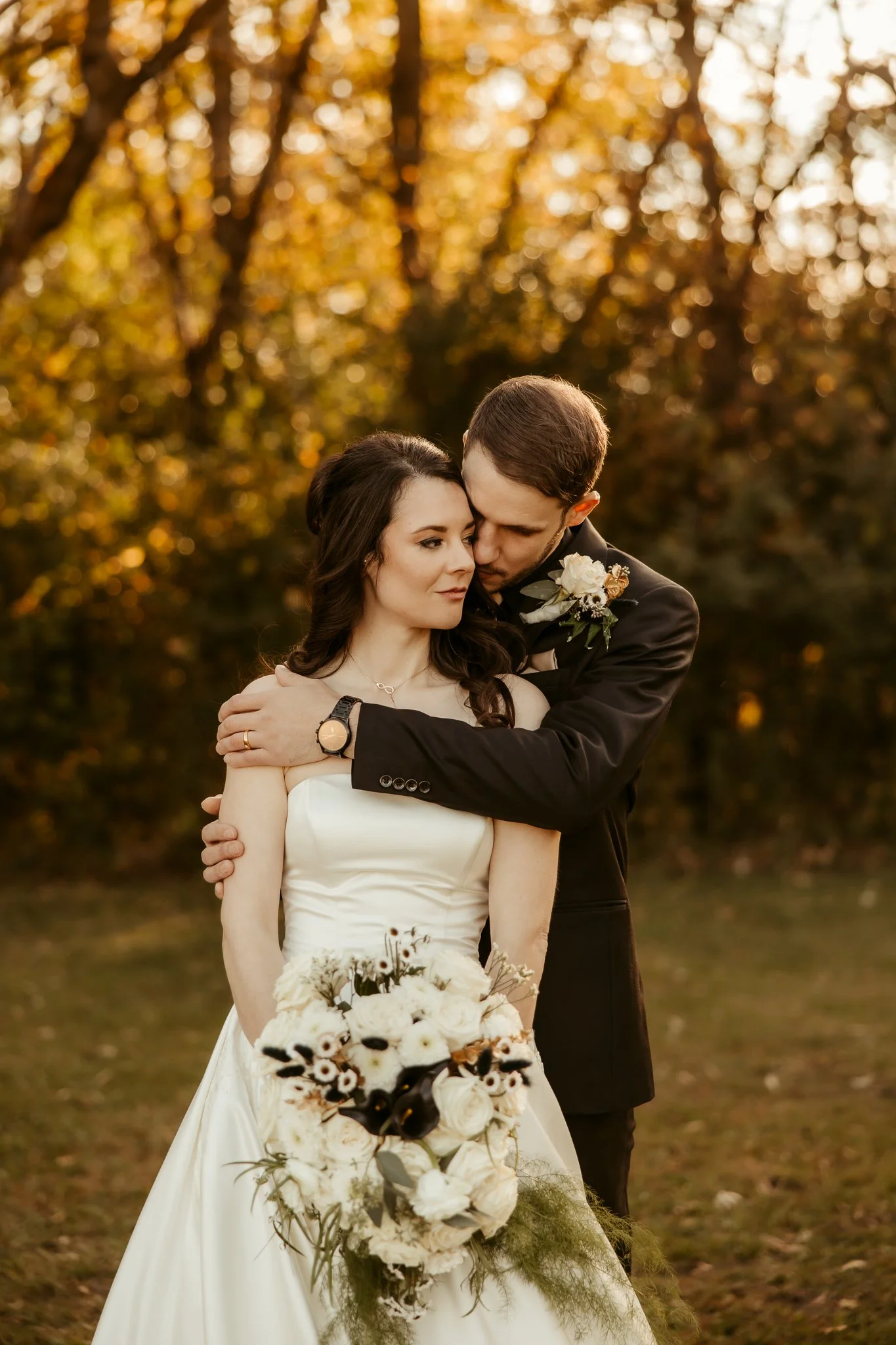 A bride and groom embracing outdoors during sunset, with autumn trees in the background. The bride has dark hair and wears a white wedding dress, holding a bouquet of white and dark flowers. The groom, with short brown hair, wears a black suit with a