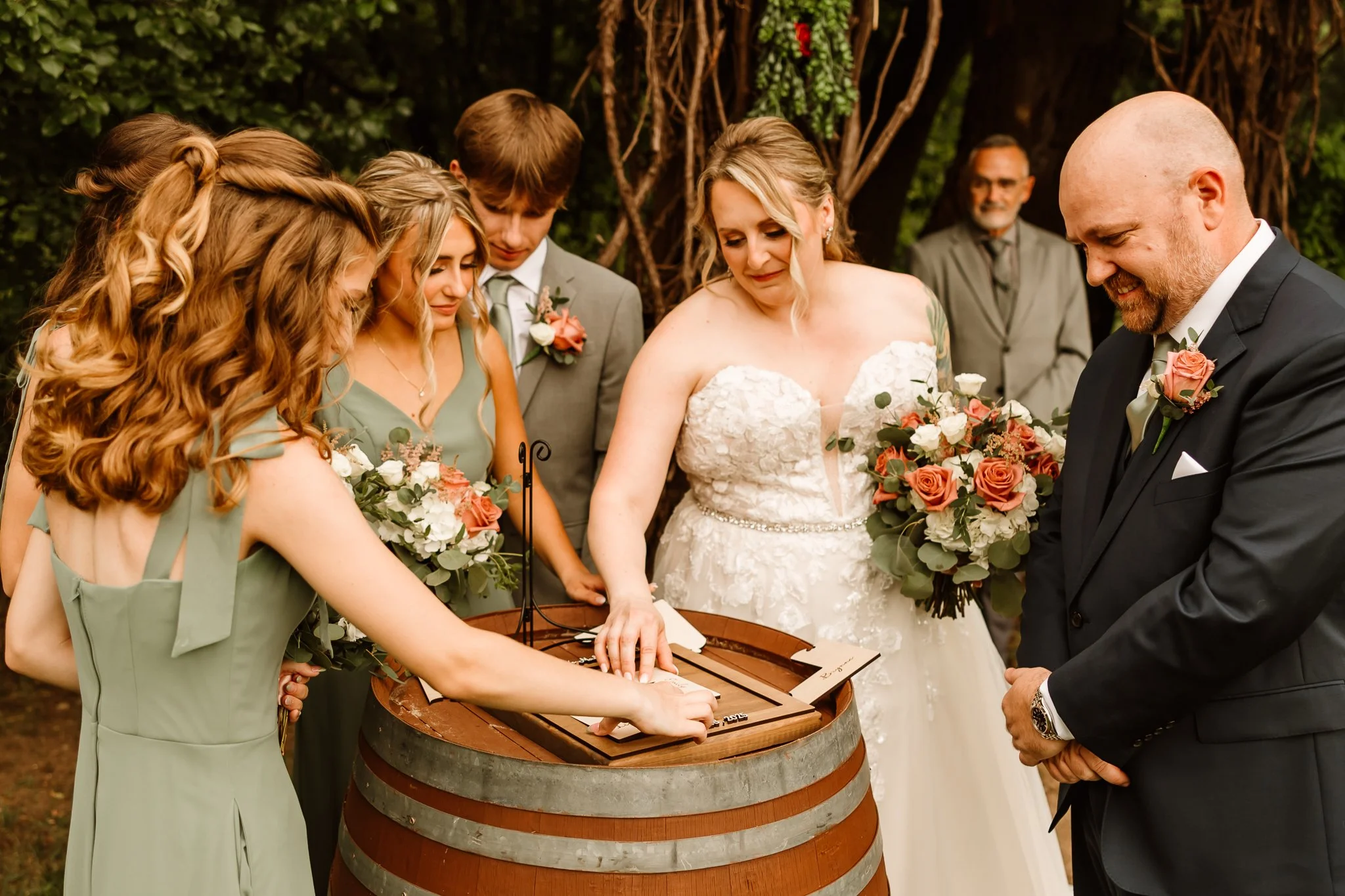Wedding ceremony with a bride, groom, and wedding party signing a document on a wooden barrel in an outdoor setting.