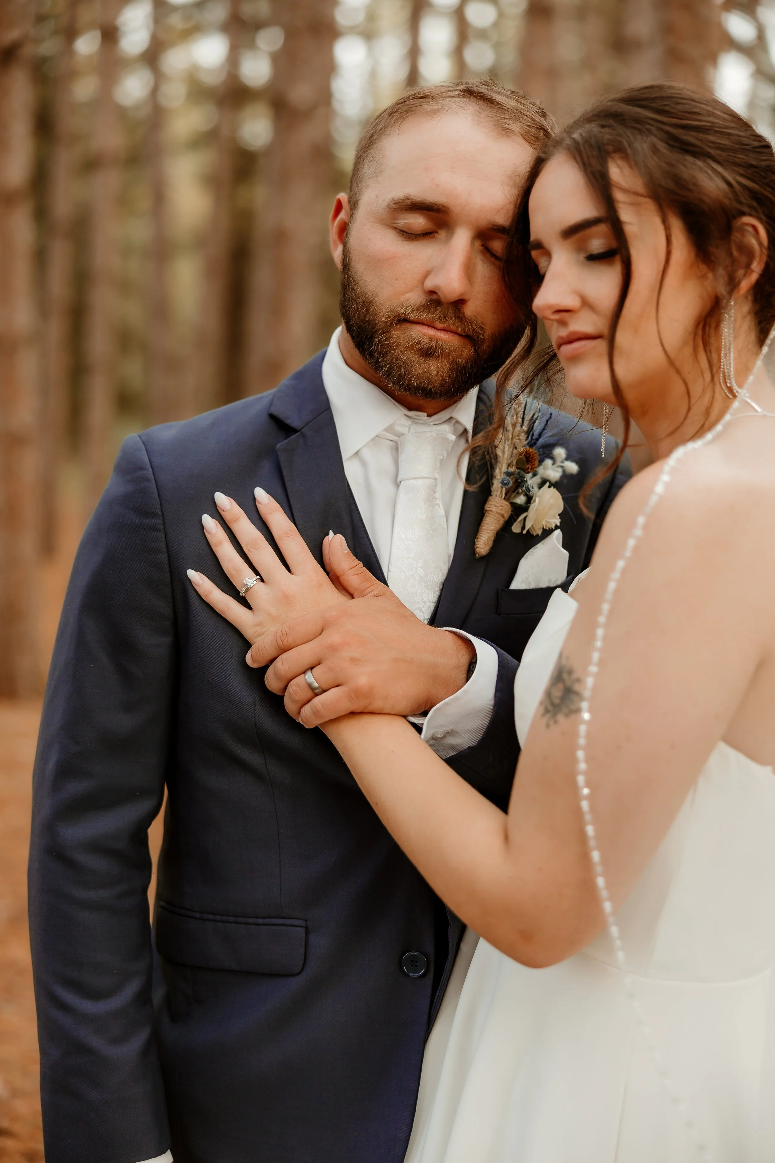 A newlywed couple, the groom in a navy suit and white tie, and the bride in a white wedding dress, standing in a forest, with their heads touching and eyes closed, holding hands.