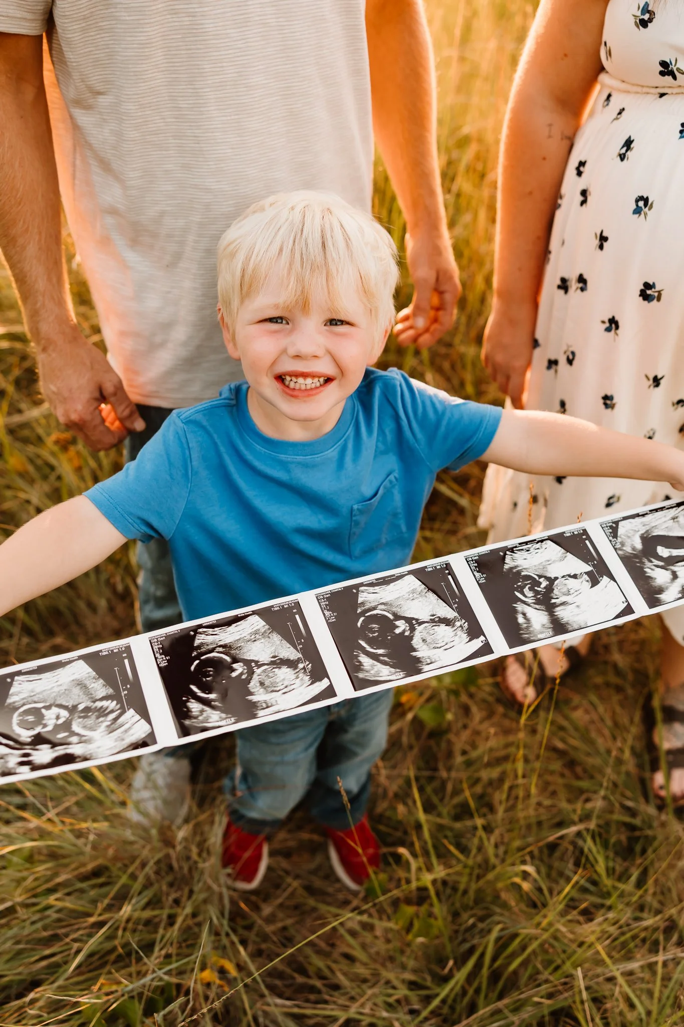 A young boy holding an ultrasound strip, surrounded by two adults, outdoors in a field during sunset, with a big smile.