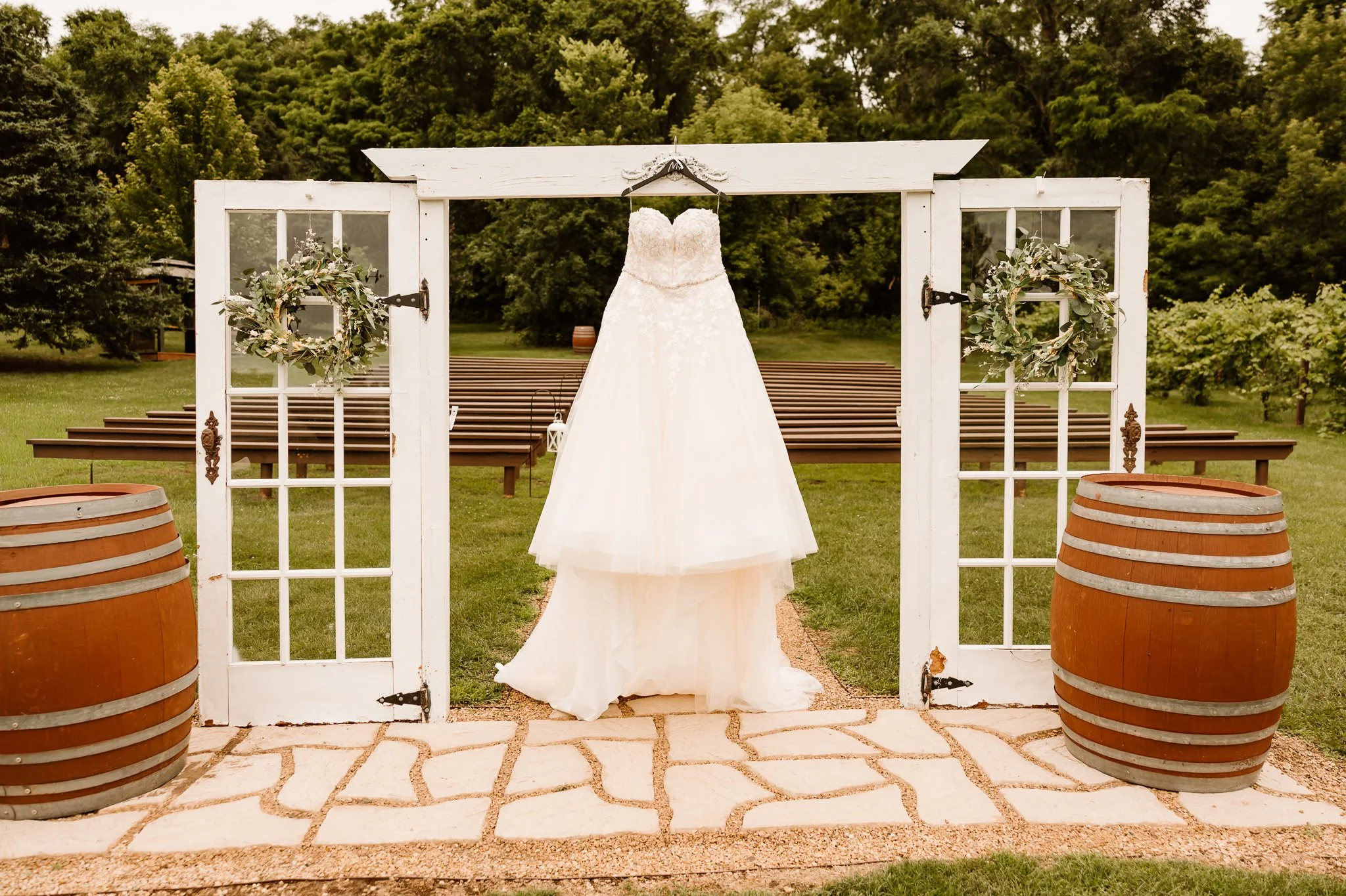 Wedding dress hanging on a wooden and glass door arch at an outdoor garden venue with trees in the background.
