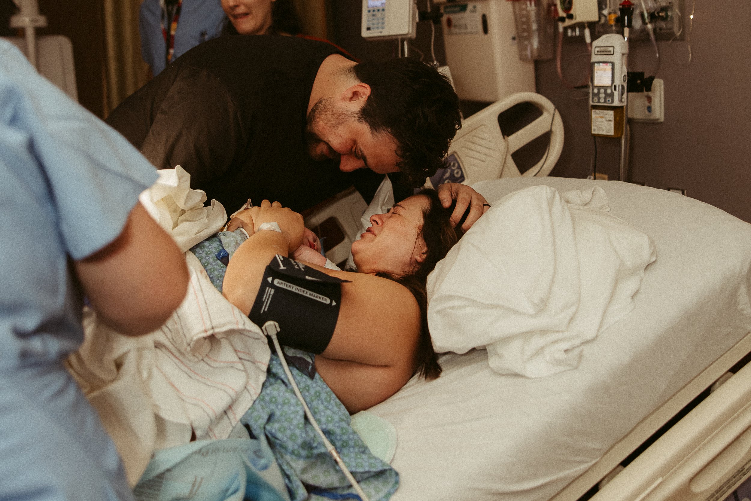 A woman lying in a hospital bed holding a newborn baby, with a man leaning over and touching her head, in a hospital room with medical equipment in the background.