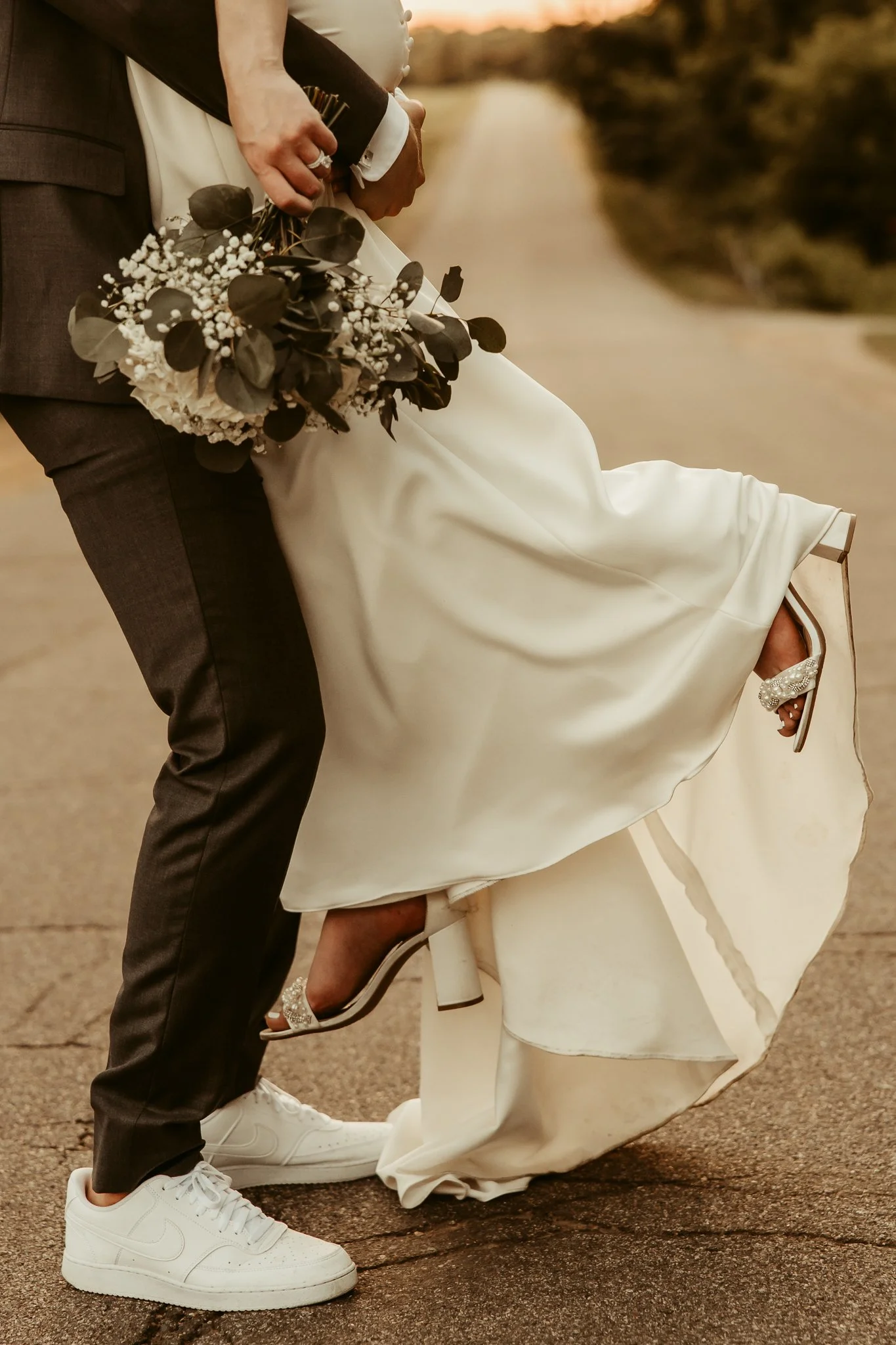 Close-up of a bride and groom, focusing on the bride's lifted dress showing her high heels and the groom's sneakers, with the bride holding a bouquet of white and green flowers, during a wedding photograph on a road at sunset.
