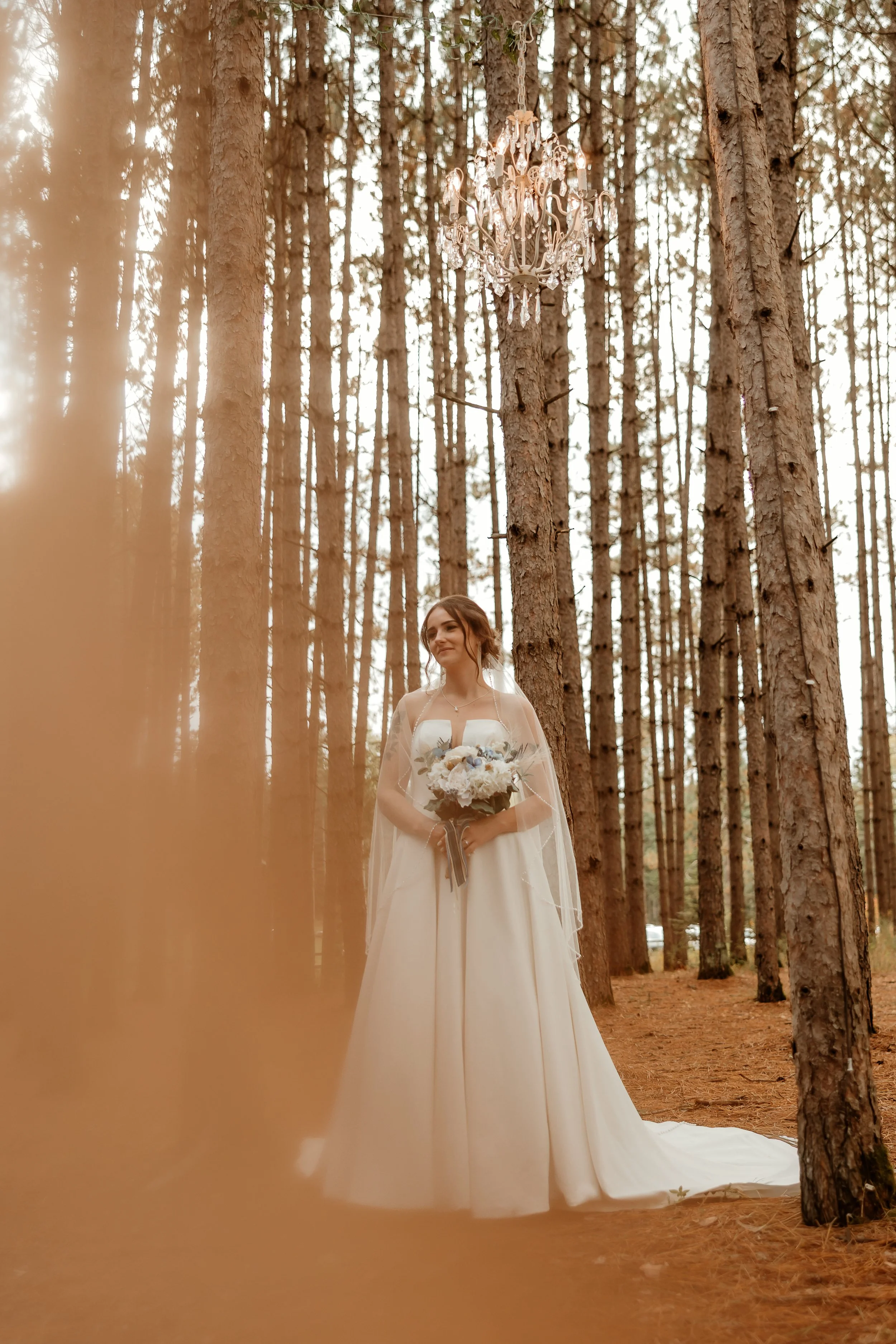A bride in a white wedding gown holding a bouquet of flowers, standing in a forest with tall pine trees, and a chandelier hanging from one of the trees.