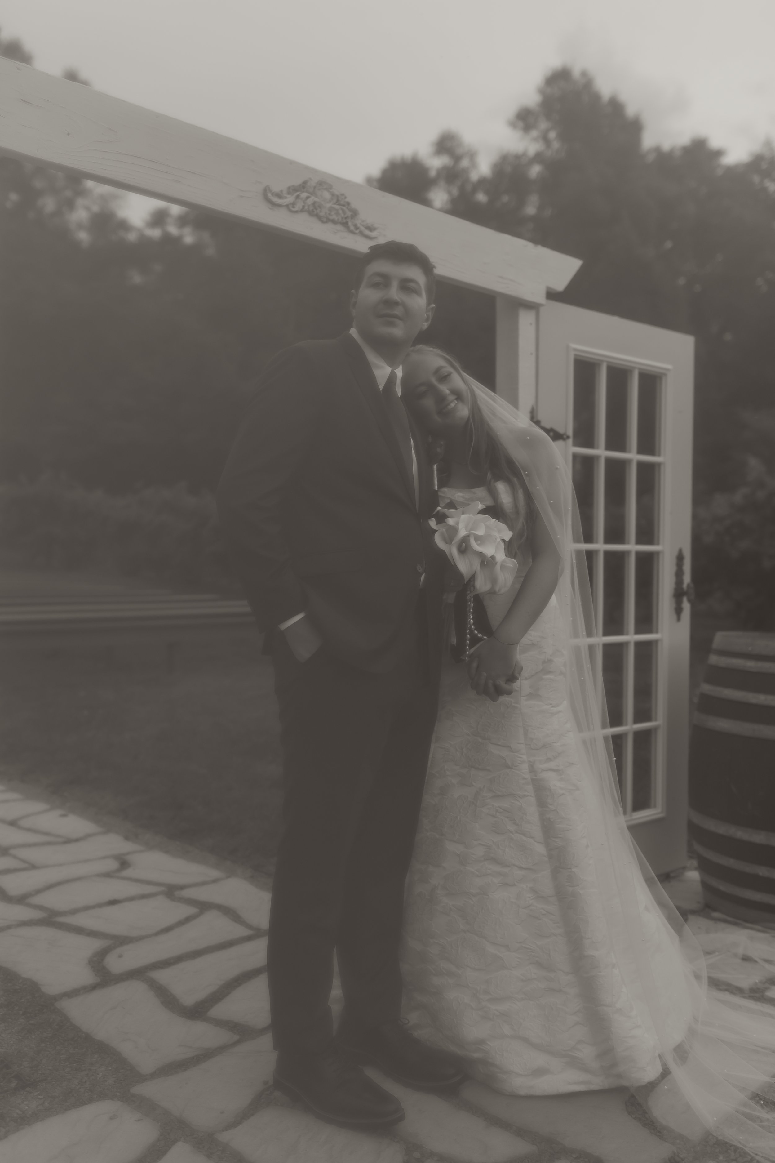 A black and white photo of a bride and groom standing outdoors, holding hands. The bride is leaning her head on the groom's shoulder, holding a bouquet of flowers, with a veil flowing down her back. The groom has his hands in his pockets, dressed in 