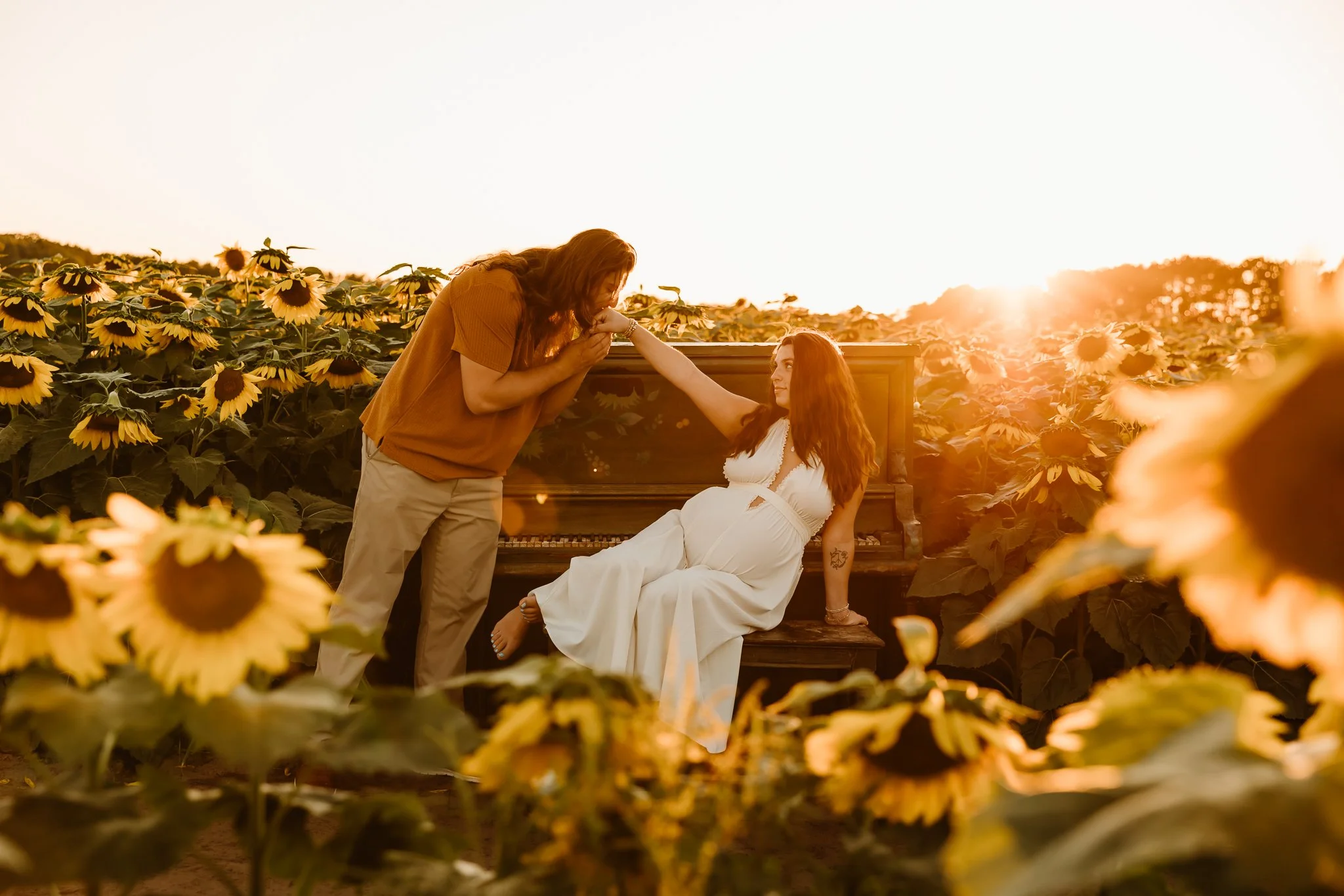A woman in a white dress sitting on a bench in a sunflower field during sunset, a man leaning over from a piano to hold her hand.