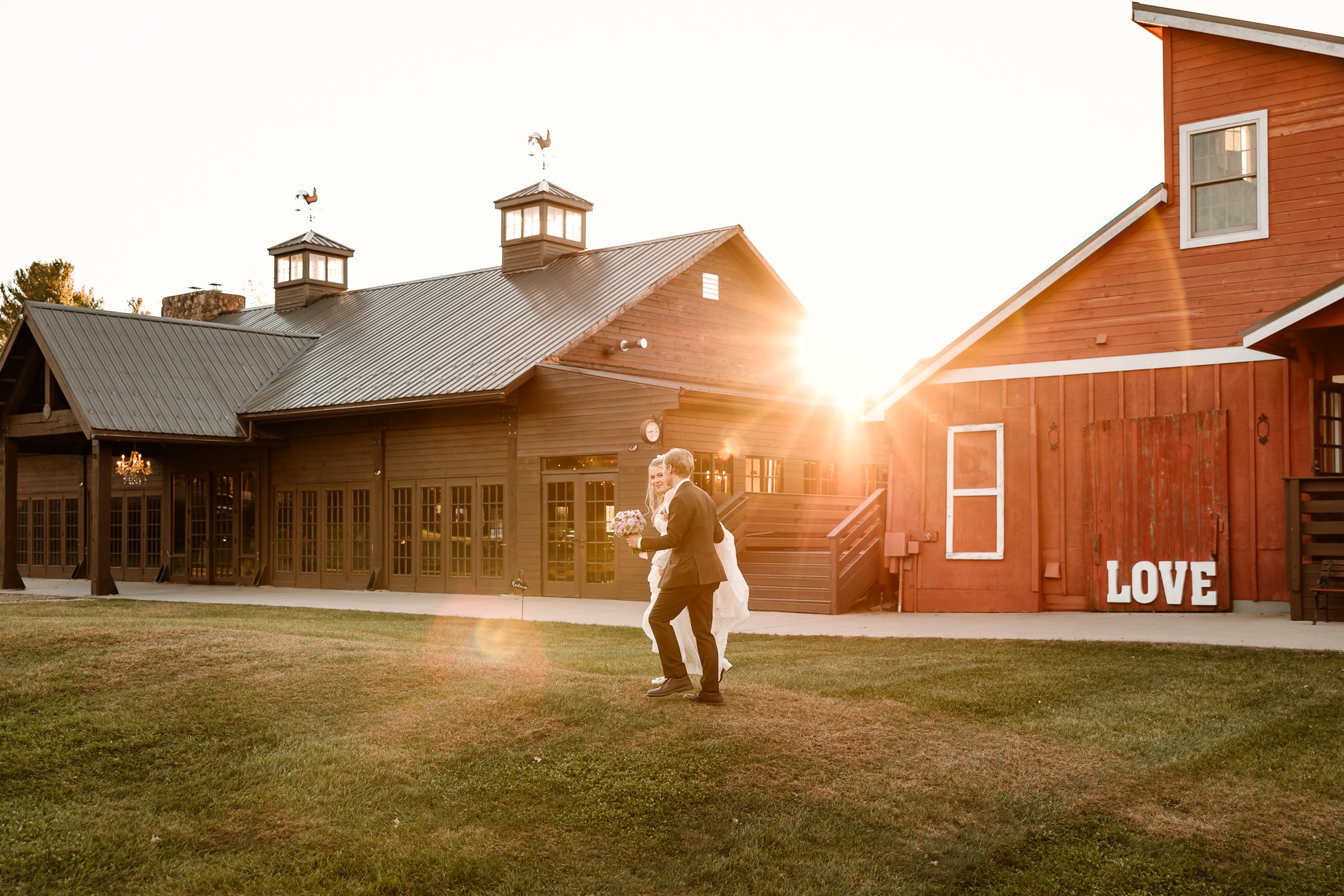 A bride and groom walking on a grassy lawn during sunset, near a rustic barn with a sign reading 'LOVE' on the side, in a rural outdoor setting.