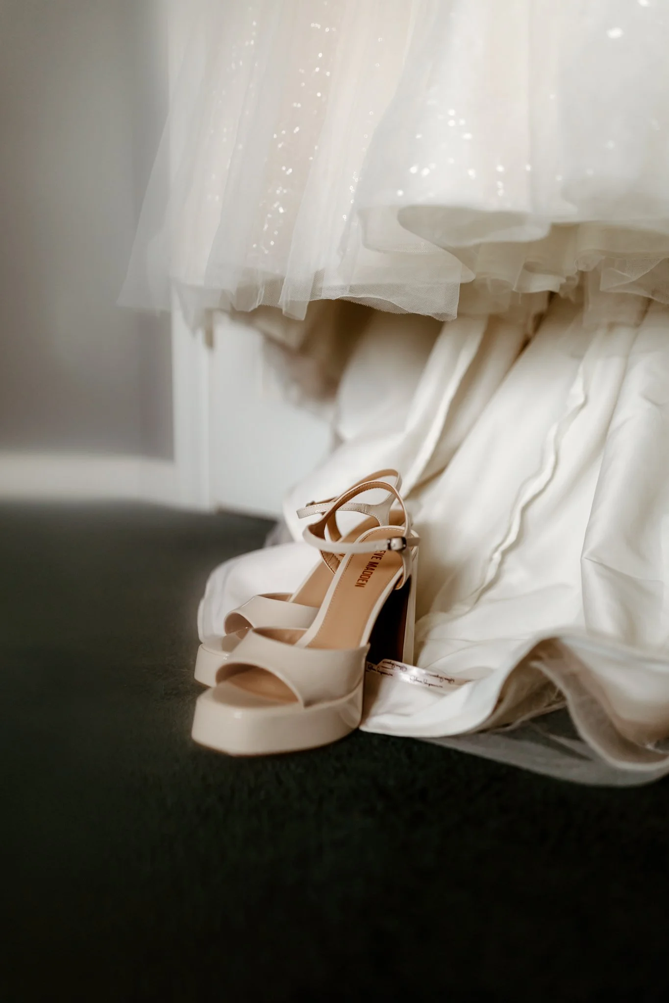 A pair of beige or nude high-heeled sandals with ankle straps resting on a dark surface, with a white wedding dress and sheer fabric in the background.