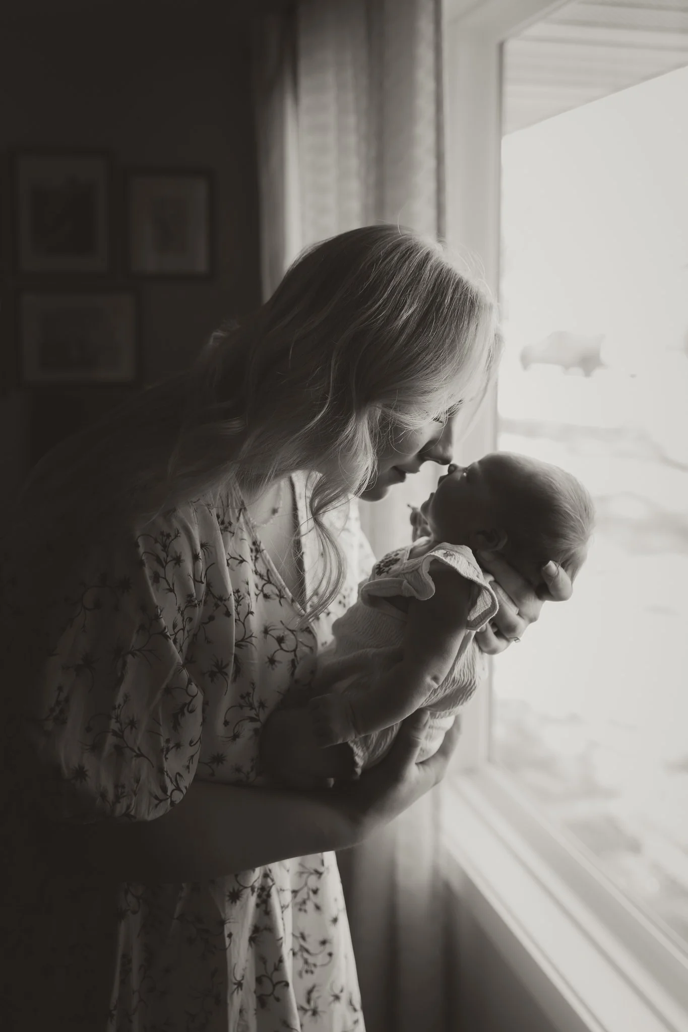 A woman holding a newborn baby near a window, with the woman gently touching noses with the baby.
