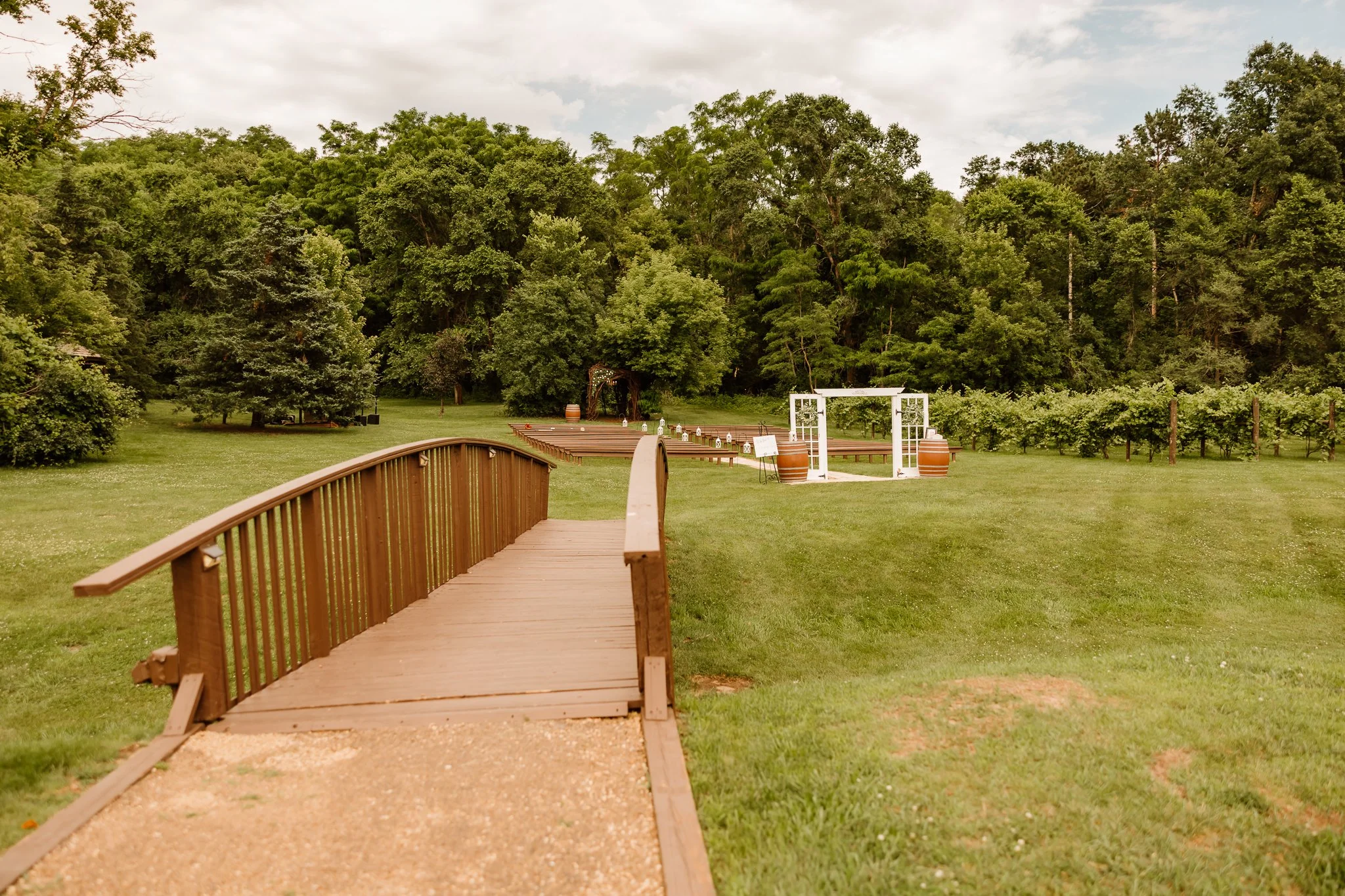 A wooden footbridge leads to a grassy area with a white wedding arch decorated with white flowers and surrounded by large wine barrel planters, set against a backdrop of dense green trees and an overcast sky.