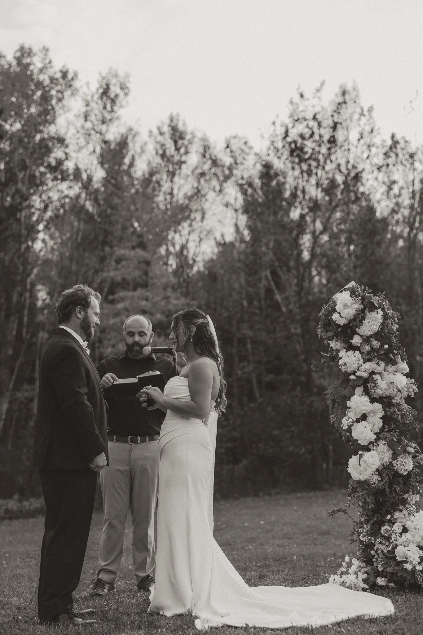 A black-and-white photo of a wedding ceremony outdoors with a bride and groom standing facing each other, a officiant holding a book and microphone, and a floral arch on a grassy area with trees in the background.