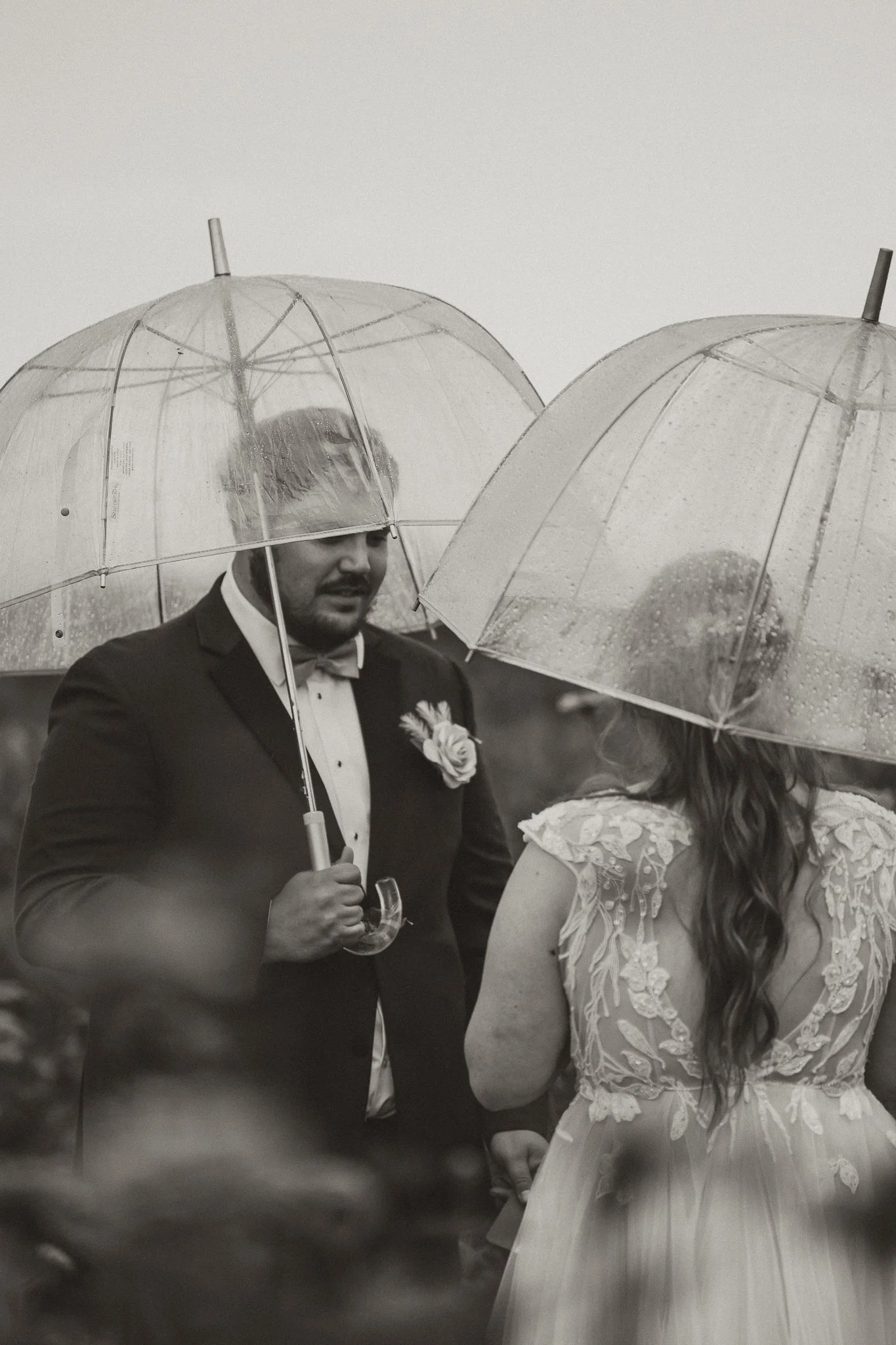 A black and white photo of a couple at a wedding holding umbrellas in the rain. The groom is wearing a tuxedo with a boutonniere, and the bride is wearing a wedding dress with lace details.