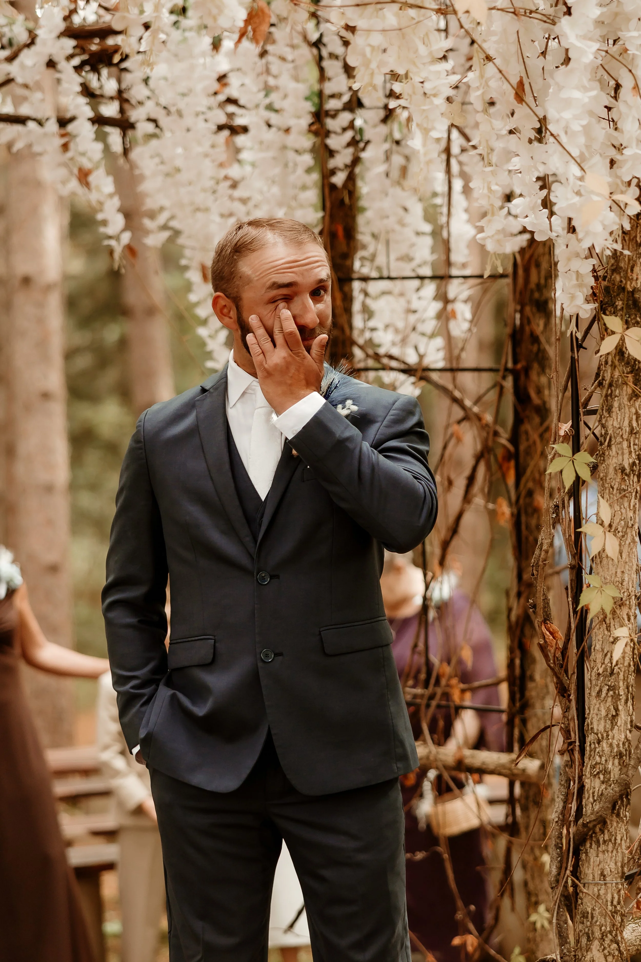 A groom in a dark suit wiping his eyes during an outdoor wedding ceremony surrounded by trees with white flowers.