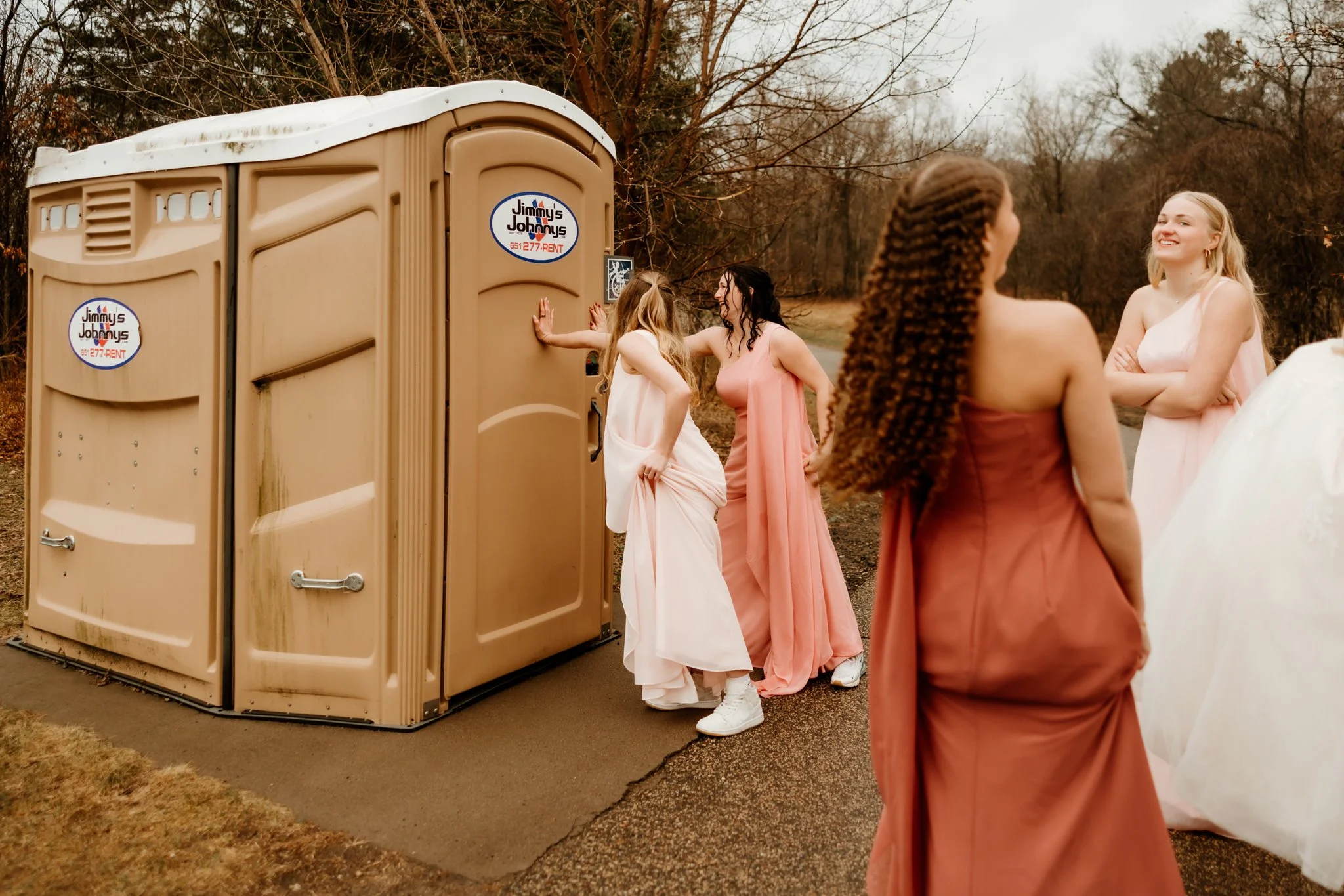Four women dressed in pink and peach dresses stand outdoors on a cloudy day near a portable toilet, engaging in conversation and smiling.