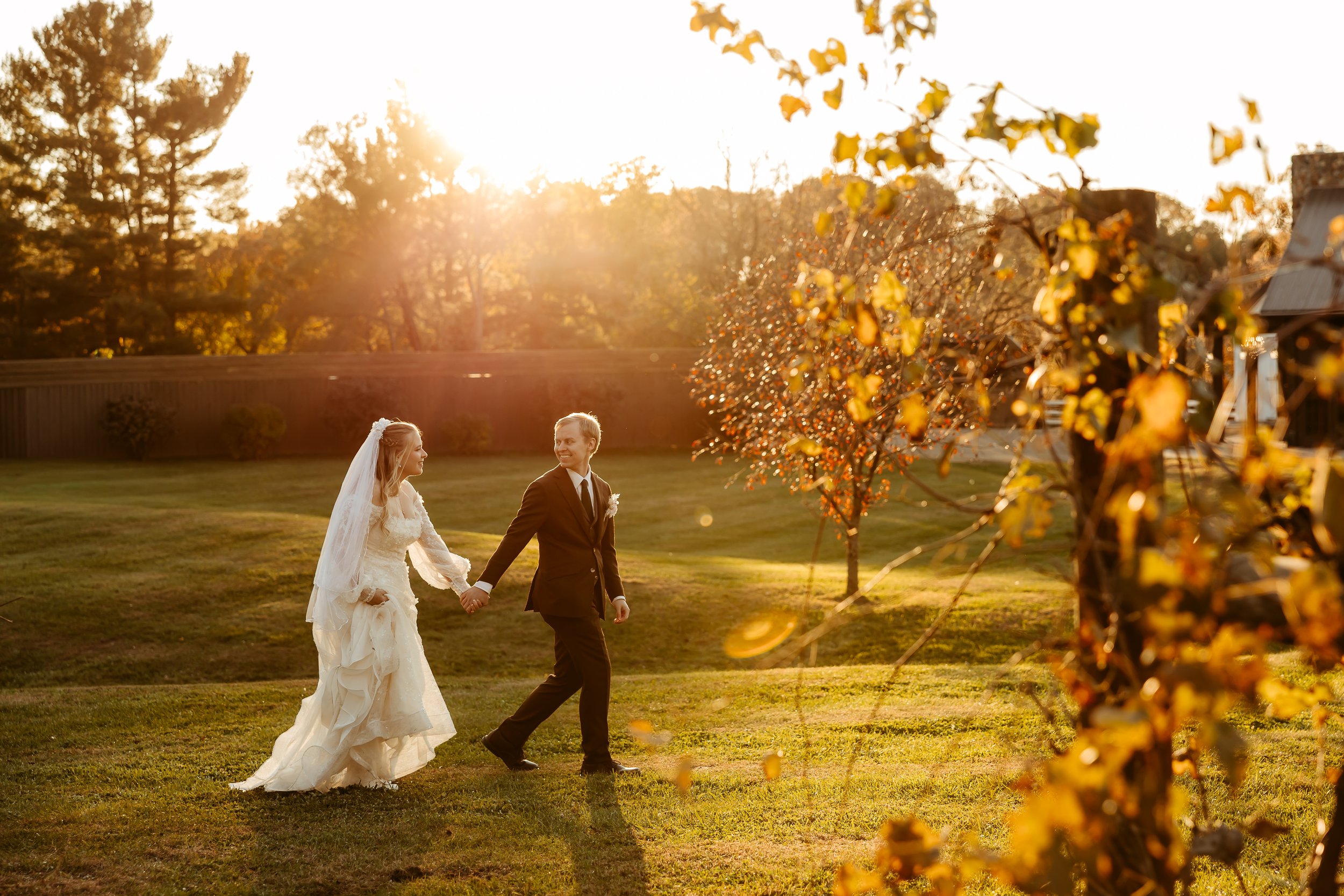 A bride and groom holding hands and walking outdoors on a sunny day, with the bride in a white wedding dress and veil, and the groom in a dark suit. The sun is setting in the background, casting a warm golden light. Minnesota wedding photographer at 