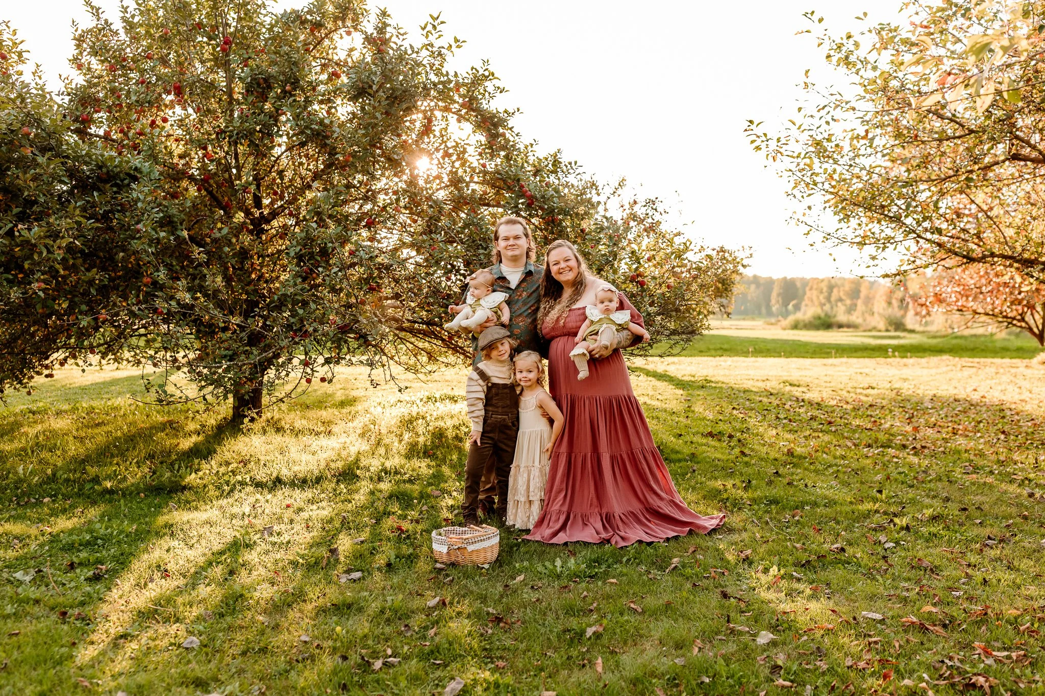 A family of five standing outdoors near apple trees during sunset. The father holds a baby girl, and the mother also holds a baby girl. Two children, a boy and a girl, stand in front, smiling. The family is dressed in casual, vintage-style clothing, 