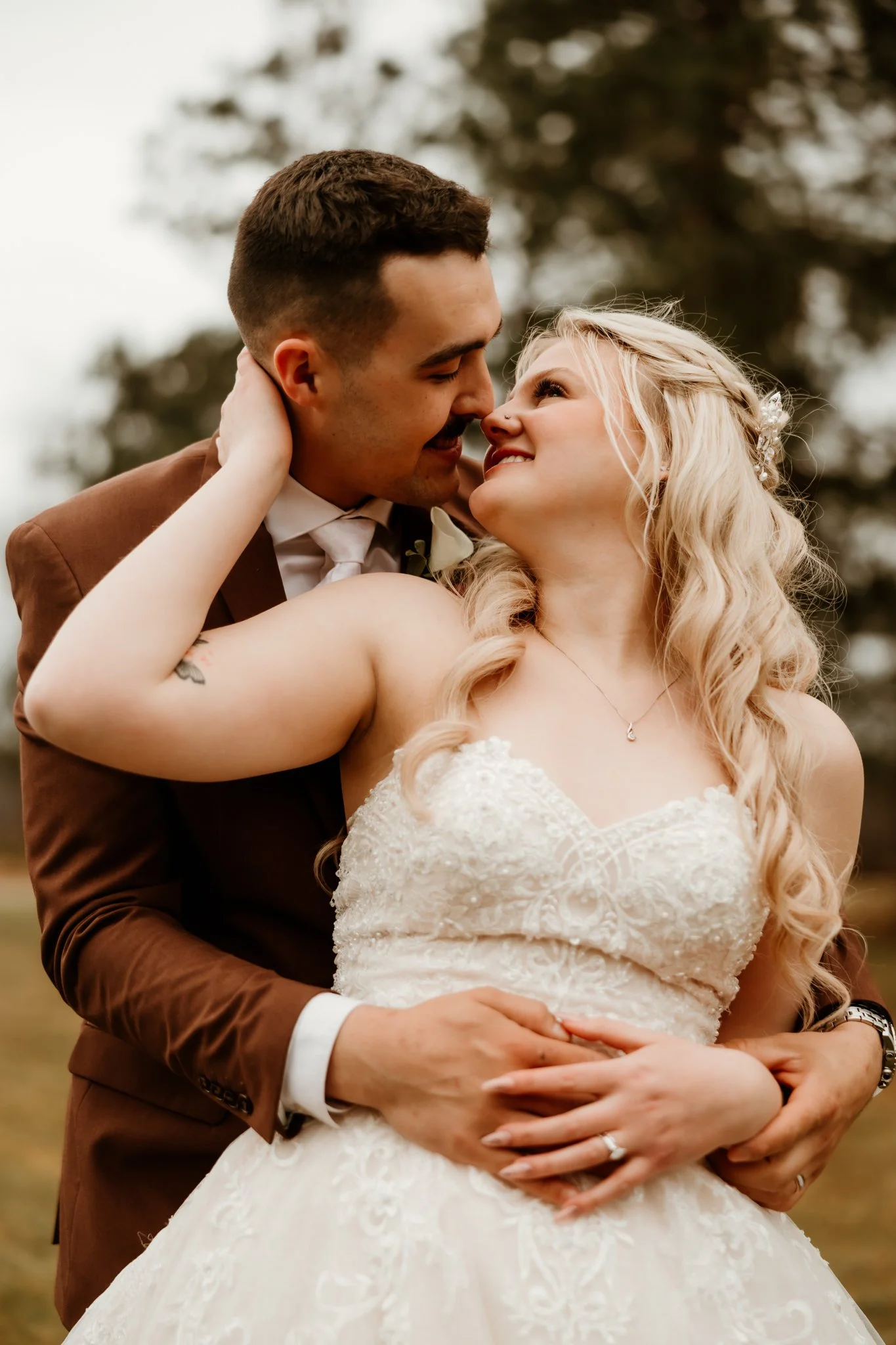 A newlywed couple shares an intimate moment outdoors, with the groom gently holding the bride and they gaze into each other's eyes, surrounded by trees.
