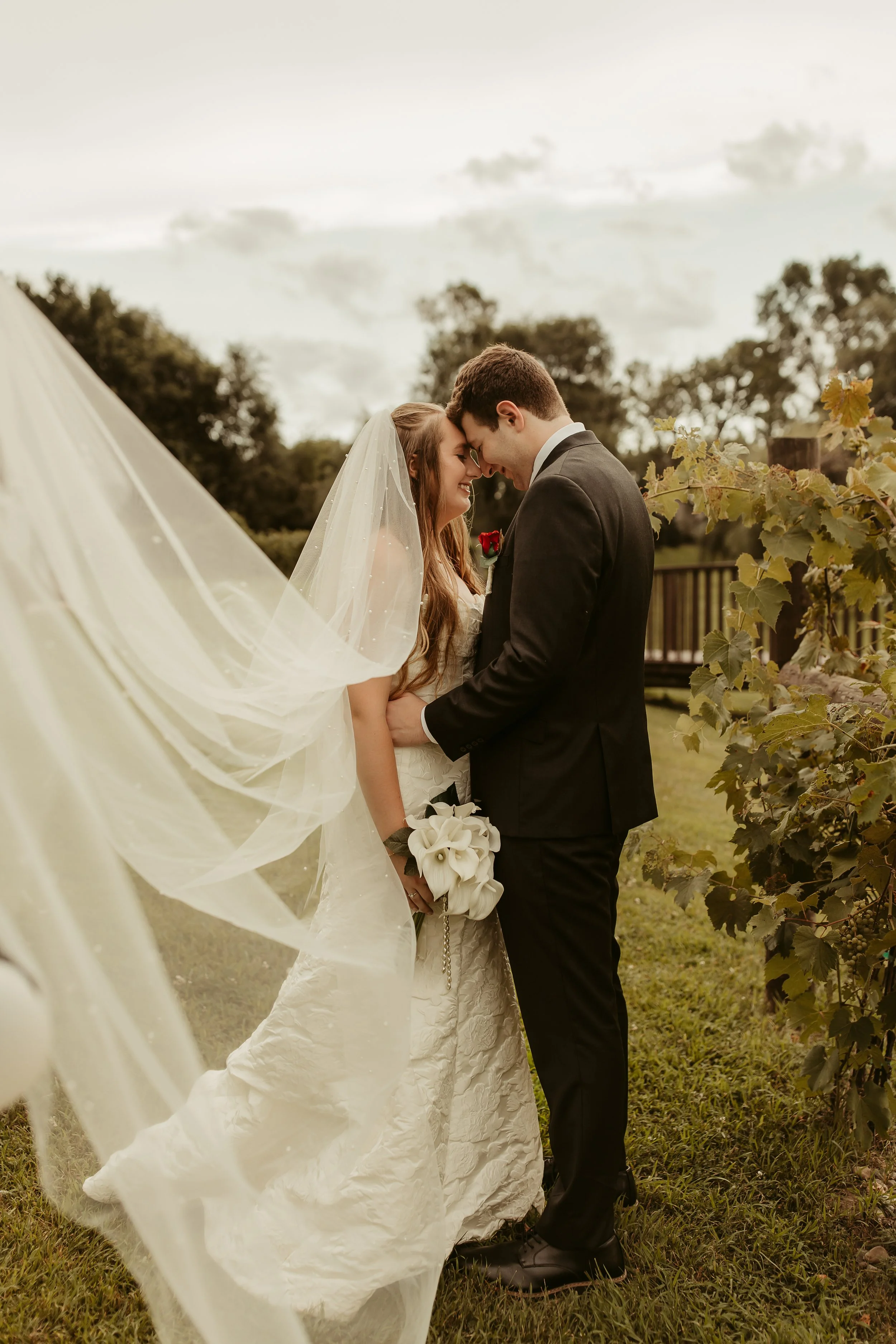 A bride and groom standing close together outdoors, touching foreheads with smiling faces, during their wedding day. The bride wears a white gown and veil, and holds a bouquet of white calla lilies. The groom wears a black suit and tie. The setting f