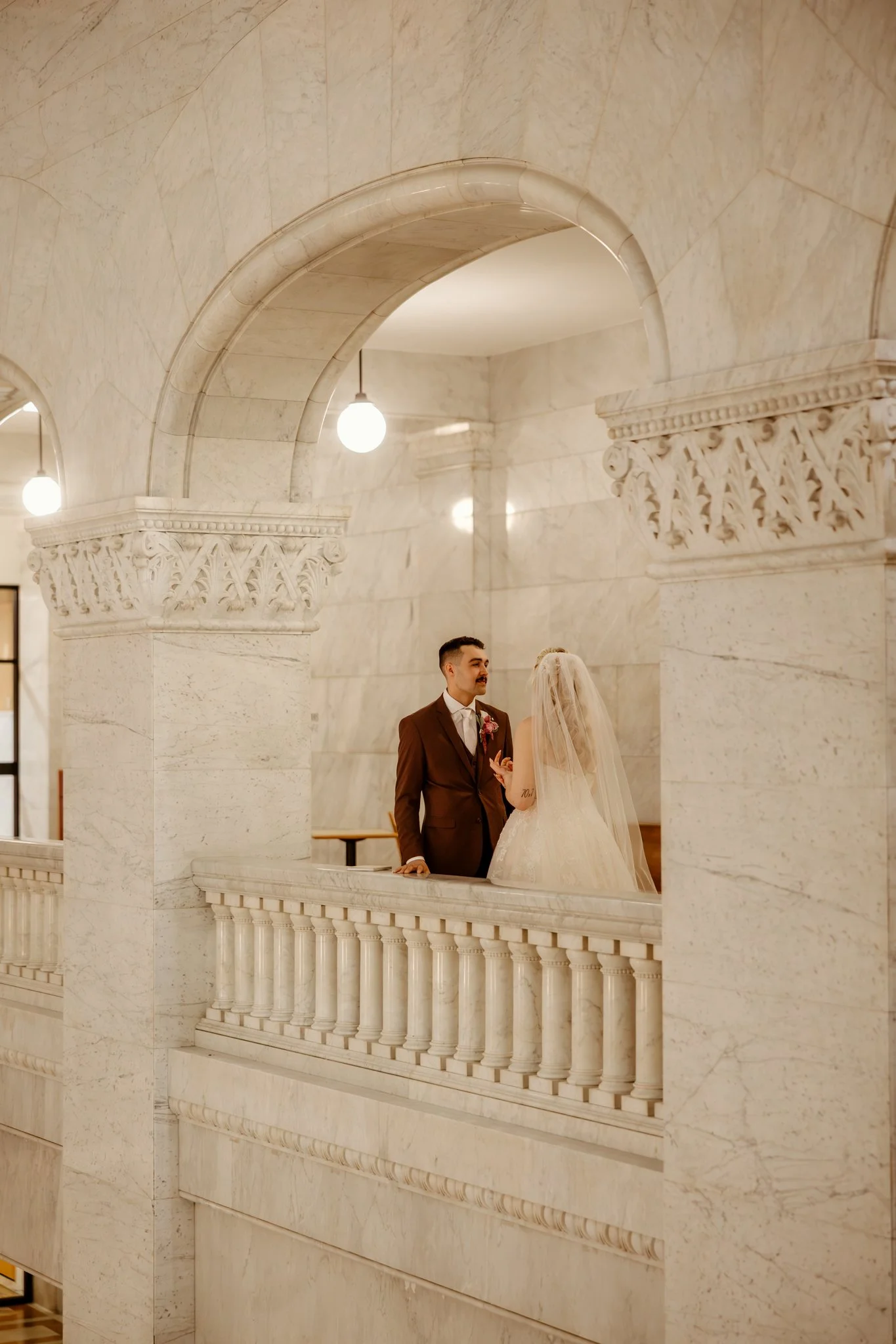 A bride and groom are standing and talking on a marble balcony inside a grand, ornate building with arches and decorative columns.