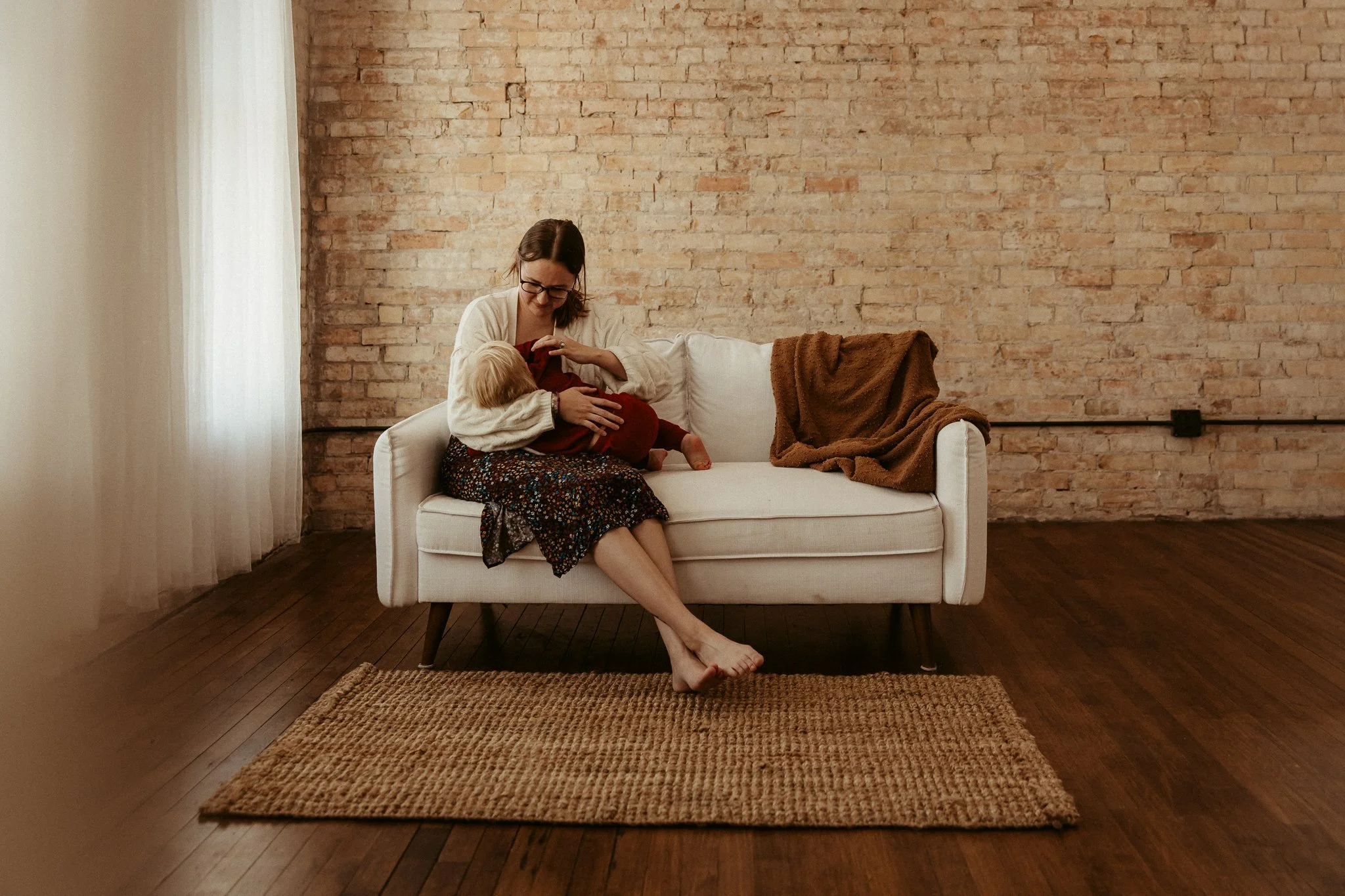 A woman breastfeeding a young child on a white sofa in a dimly lit room with wooden floors and an exposed brick wall.