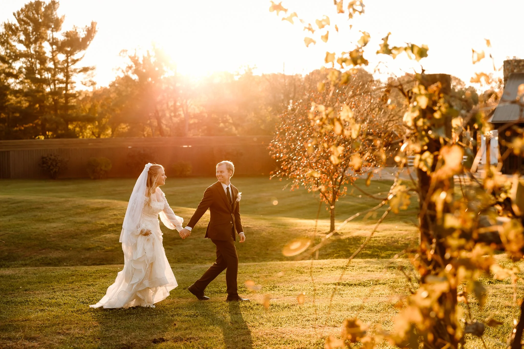 A bride and groom walking hand in hand in a grassy outdoor setting during sunset with trees and a wooden fence in the background.