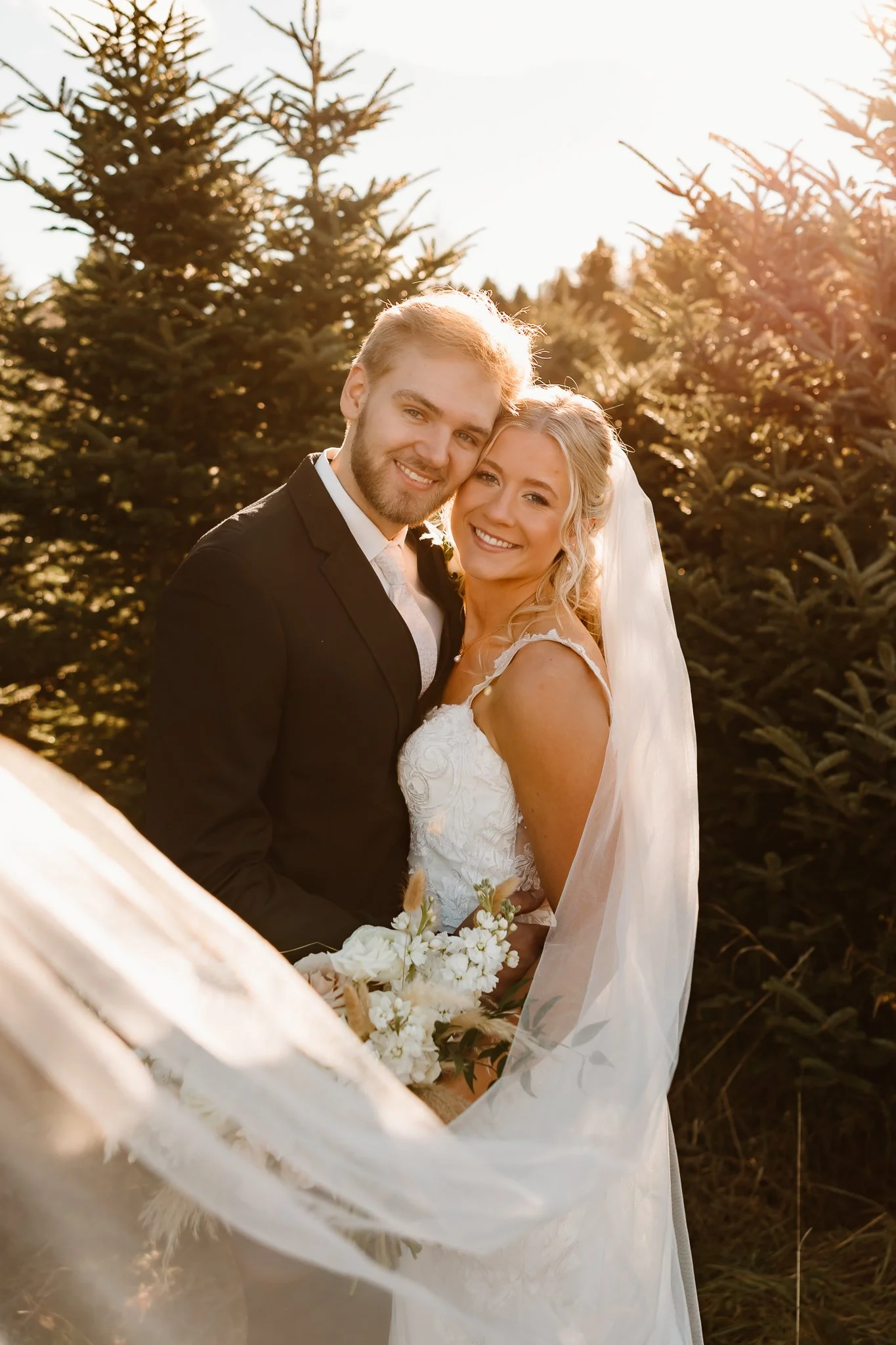 A bride and groom smiling together outdoors during sunset, with trees in the background. The bride wears a white wedding dress and veil, holding a bouquet. The groom wears a black suit and white shirt.