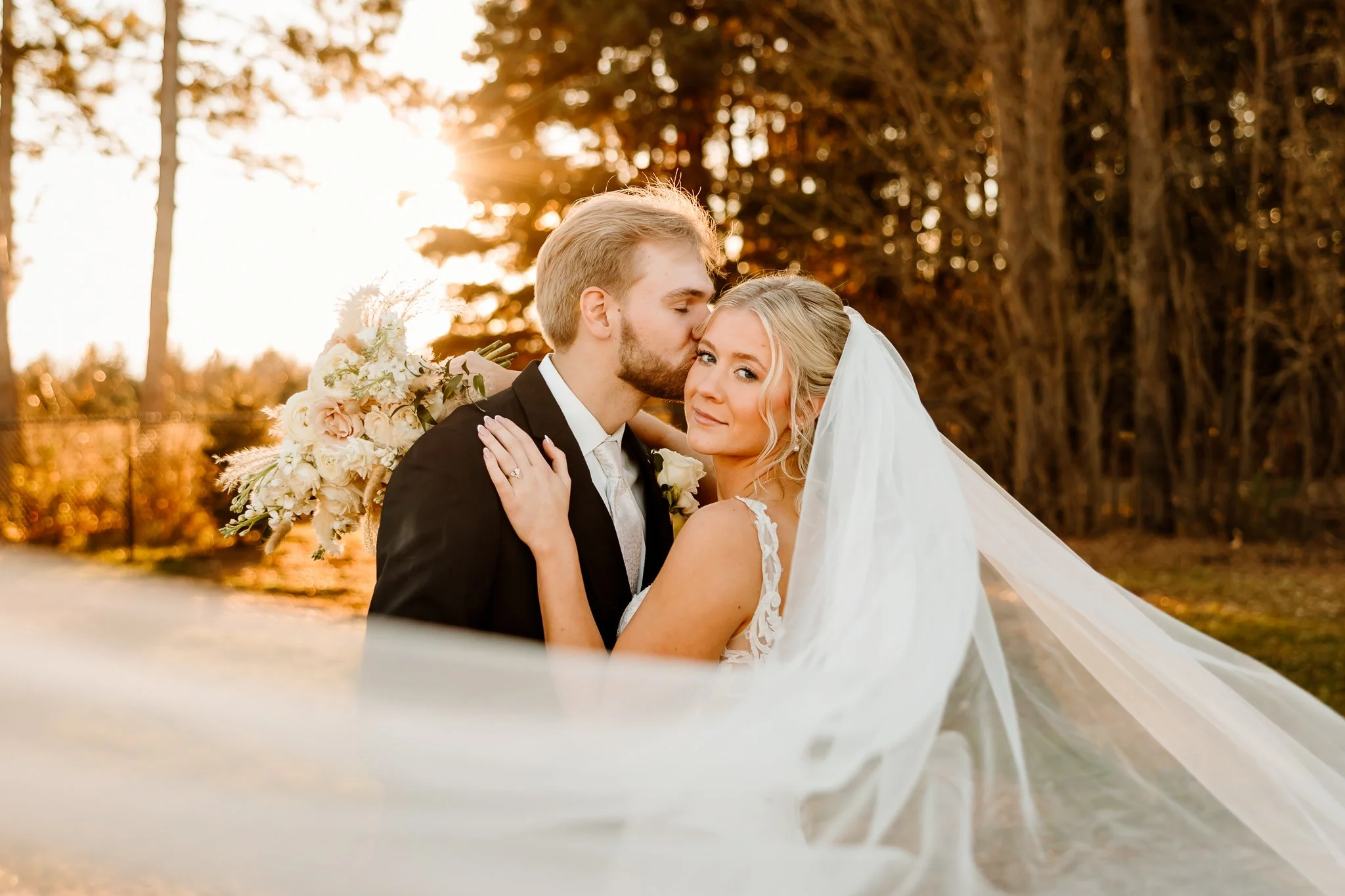 A bride and groom sharing a romantic moment outdoors during sunset, with the groom kissing the bride's temple and the bride looking at the camera, holding a bouquet of flowers, surrounded by trees. Cambridge Minnesota 