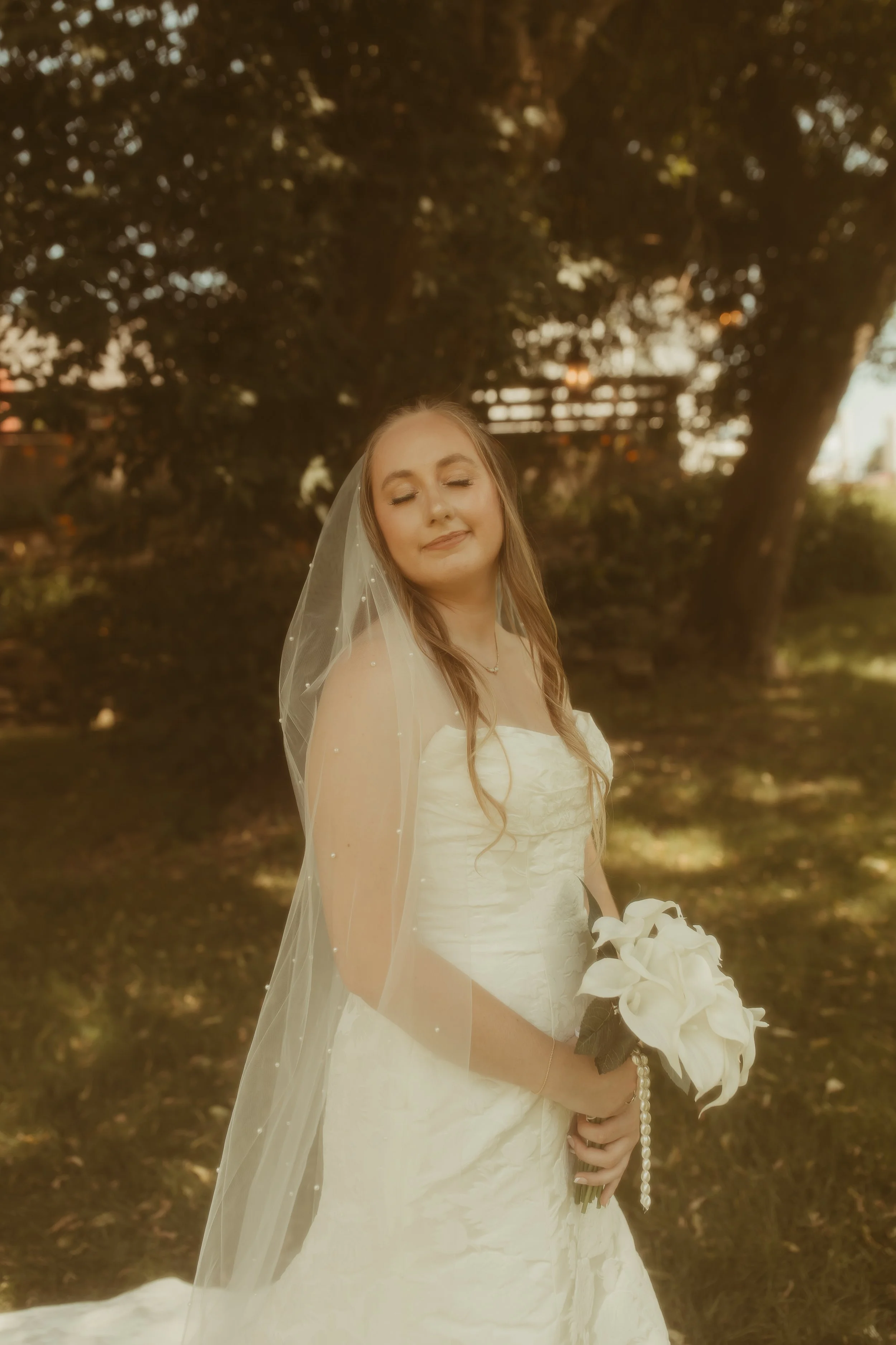 A bride in a white wedding dress holding a bouquet of white lilies, standing outdoors with trees in the background.