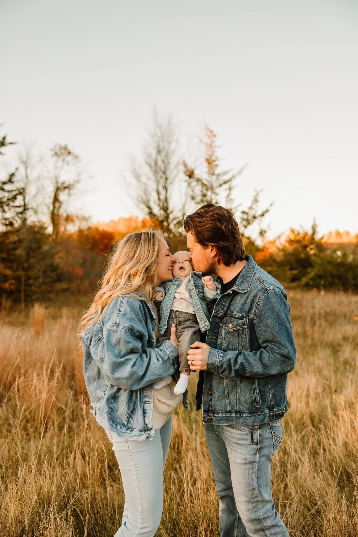 A family of three, a man, a woman, and a young child, standing in a field with trees in the background during sunset. The woman and man are holding the child between them, touching foreheads, with the woman smiling and the man kissing the child on th