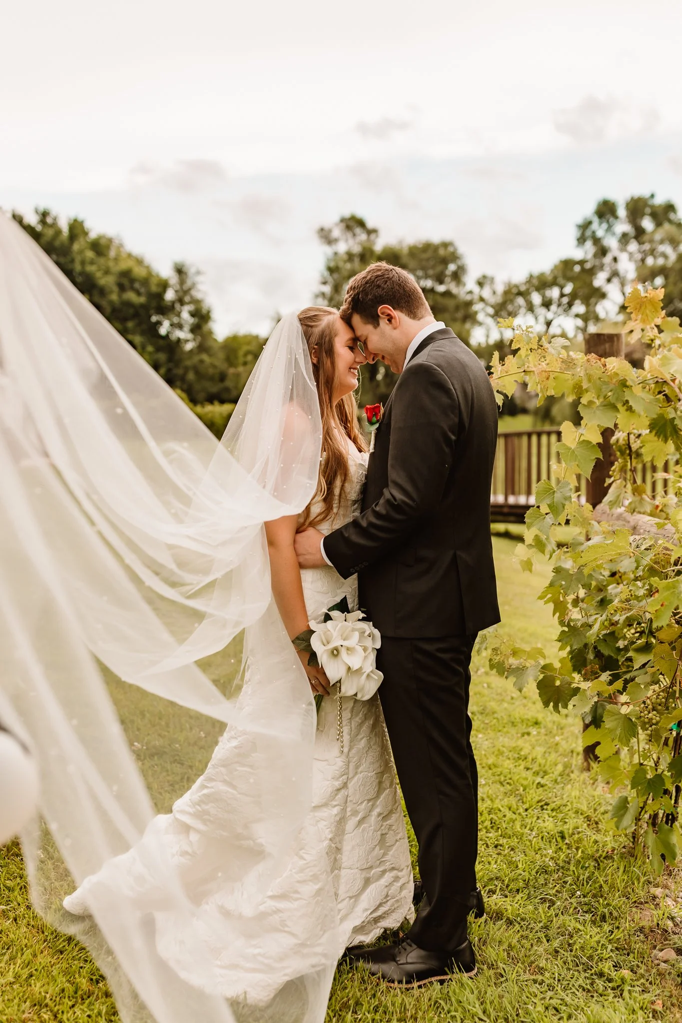 A bride and groom sharing a kiss outdoors during their wedding, with the bride holding a bouquet of white calla lilies and wearing a white wedding gown with a veil, surrounded by greenery and a cloudy sky.