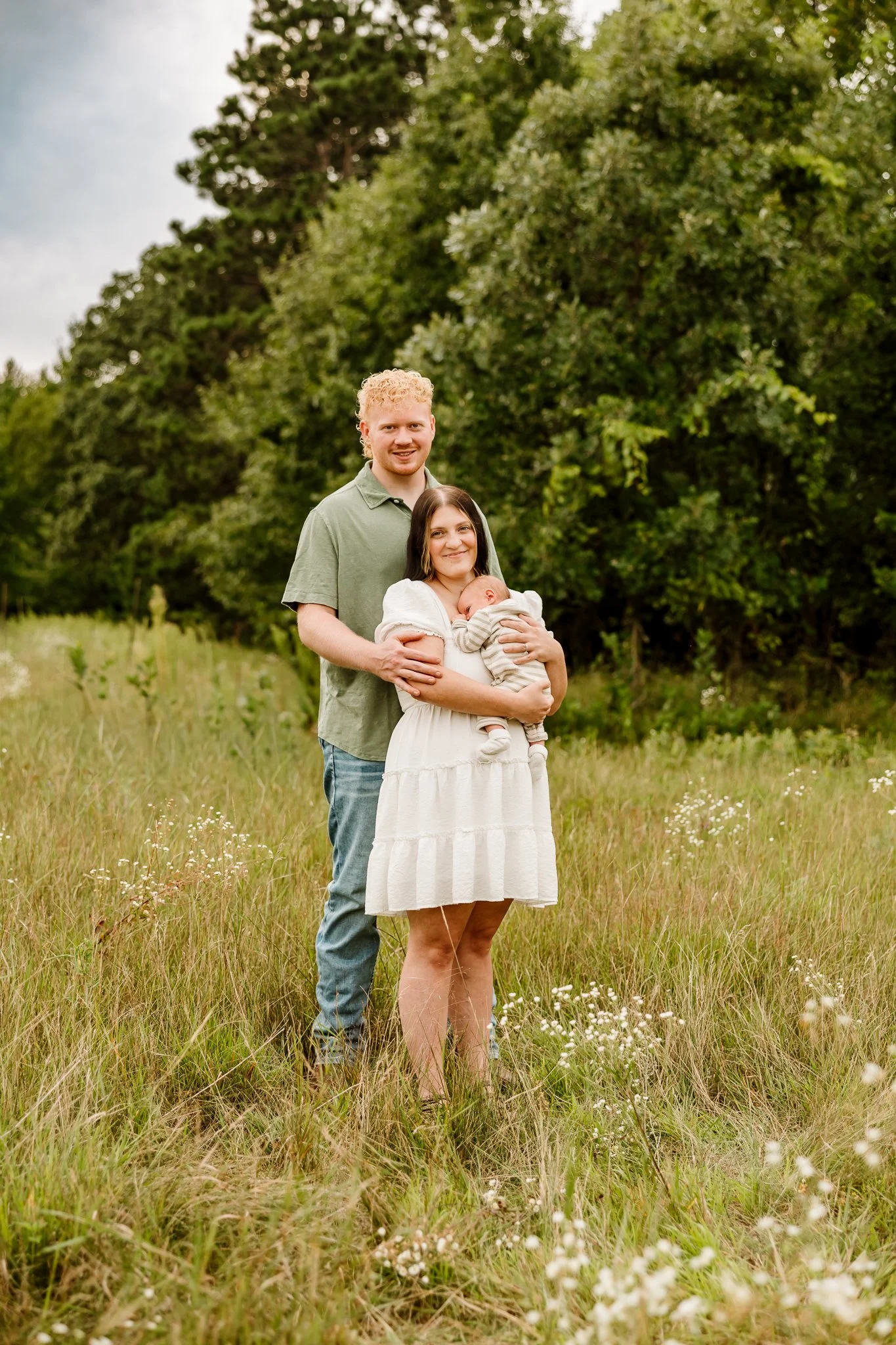 A family of three, including a woman holding a baby and a man standing behind her, standing in a grassy field with trees in the background.