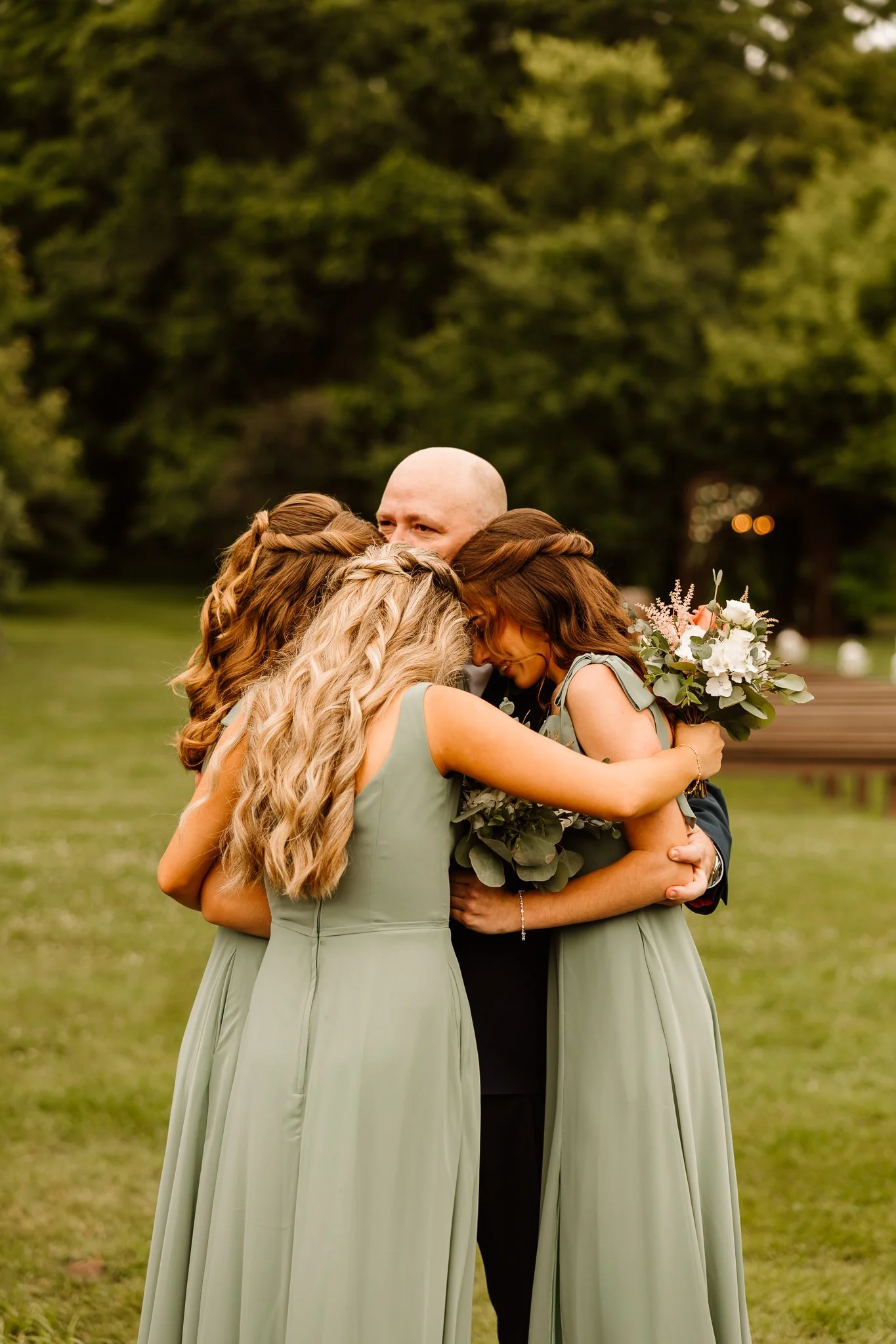 A group of four people, two women in green dresses, a man in a suit, and a woman holding a bouquet, are hugging each other outdoors in a park with green trees in the background.