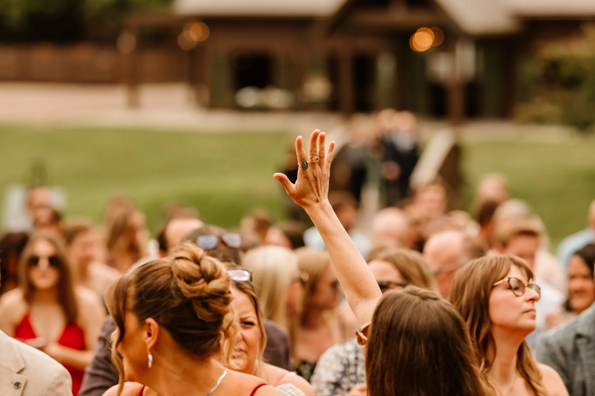 A woman raising her hand among a crowd of people at an outdoor event, with a blurred background of a building and greenery.