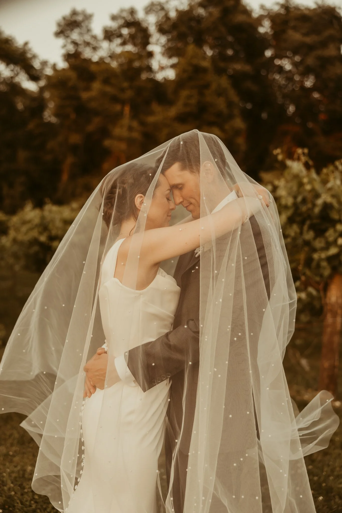 A bride and groom are embracing under a veil during their outdoor wedding at sunset.