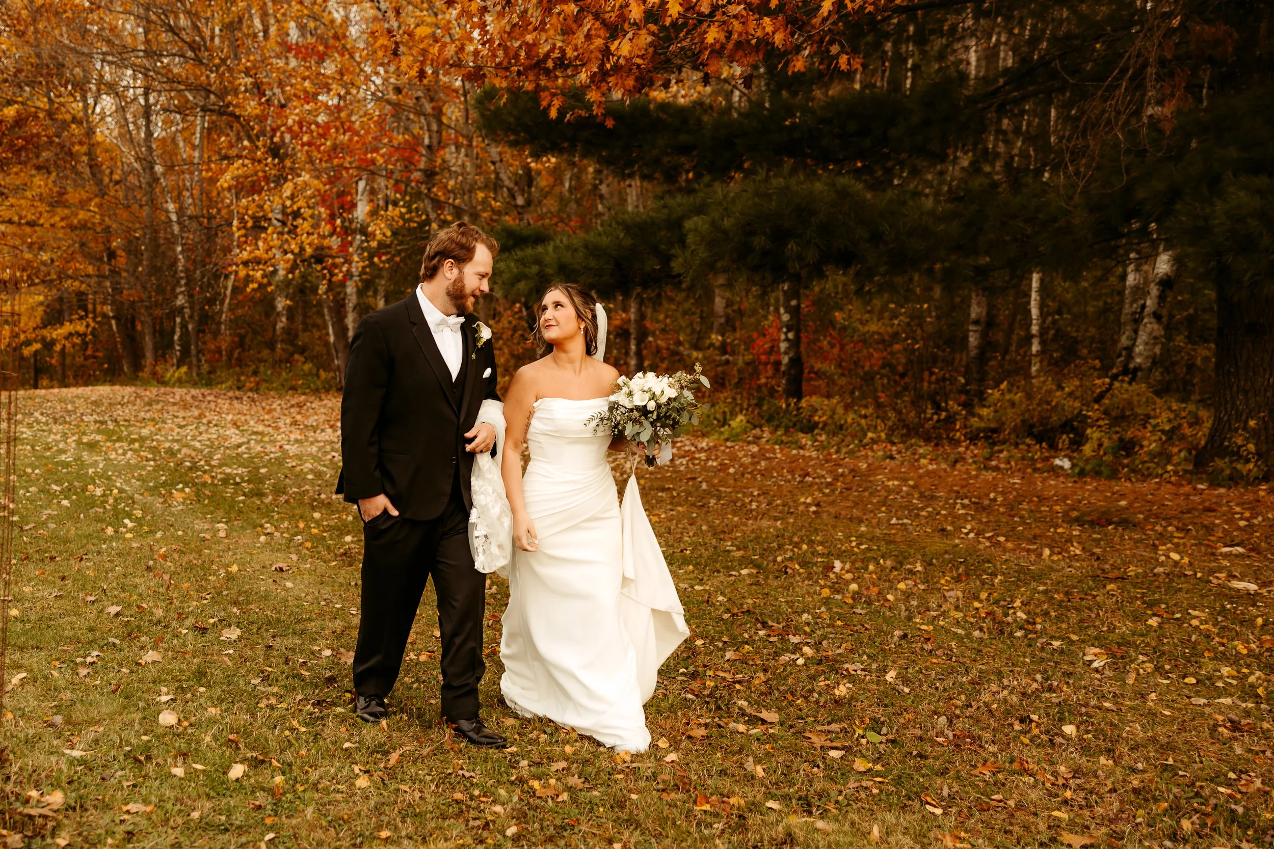 A bride and groom walk arm-in-arm through a park with autumn foliage. The bride wears a white strapless wedding gown and holds a bouquet, while the groom wears a black tuxedo with a white shirt and bow tie. They share a joyful moment. minnesota  wedd