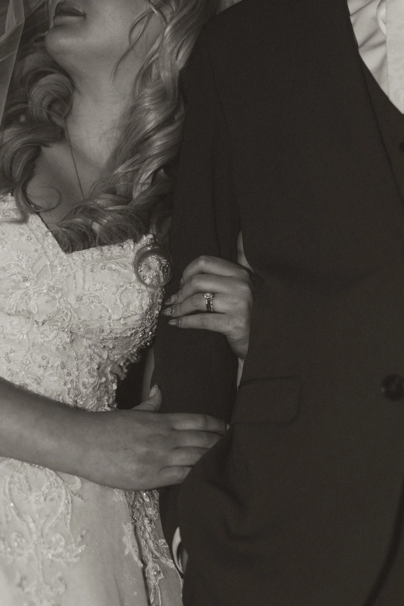 Close-up of a woman in an ornate lace wedding dress with curled hair, holding hands with a man in a dark suit, showing a wedding ring.